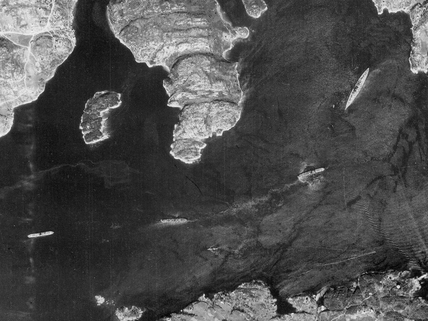A historic black and white vertical aerial photo of a fjord. A large battleship is at anchor at the right-hand end. 3 smaller ships are spread out along the fjord towards the left.
