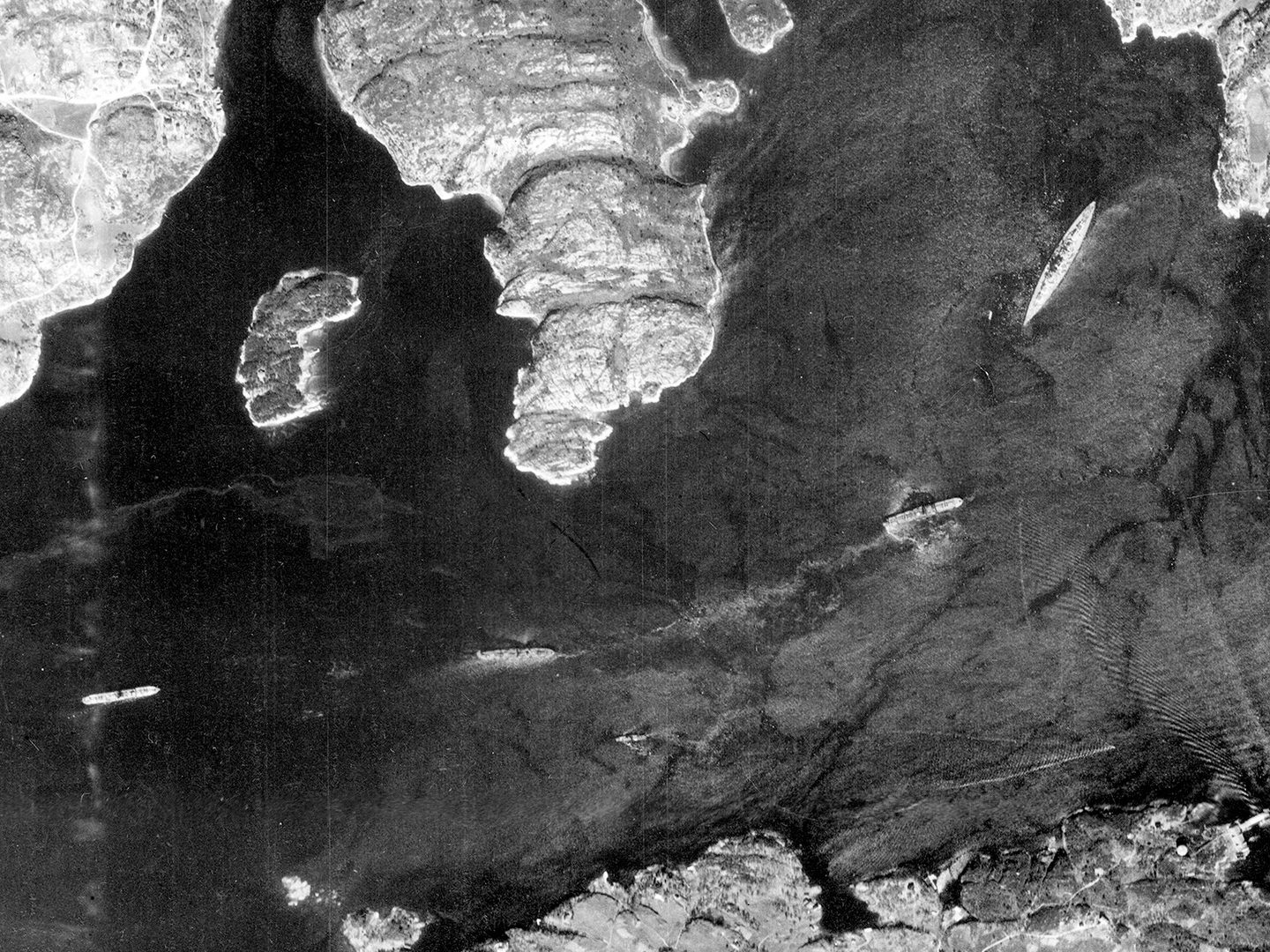 A historic black and white vertical aerial photo of a fjord. A large battleship is at anchor at the right-hand end. 3 smaller ships are spread out along the fjord towards the left.