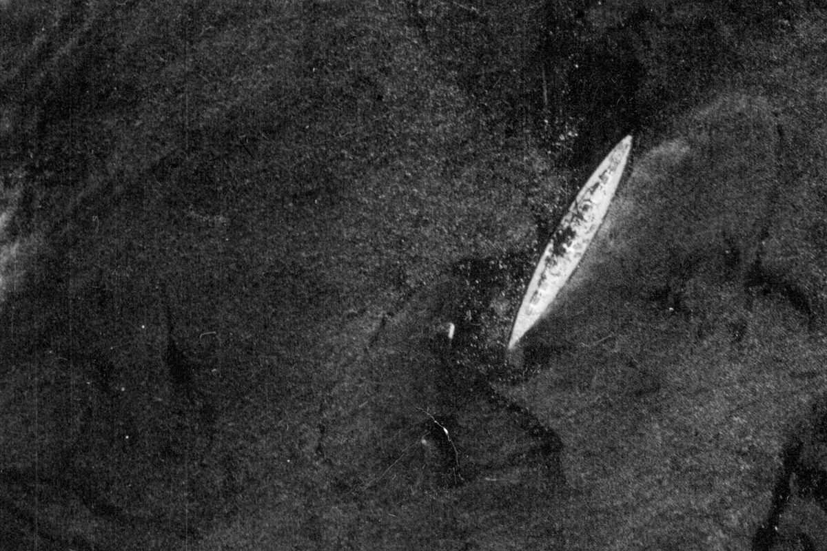 A historic black and white vertical aerial photo of the battleship Bismarck. The pale ship stands out on the dark sea.