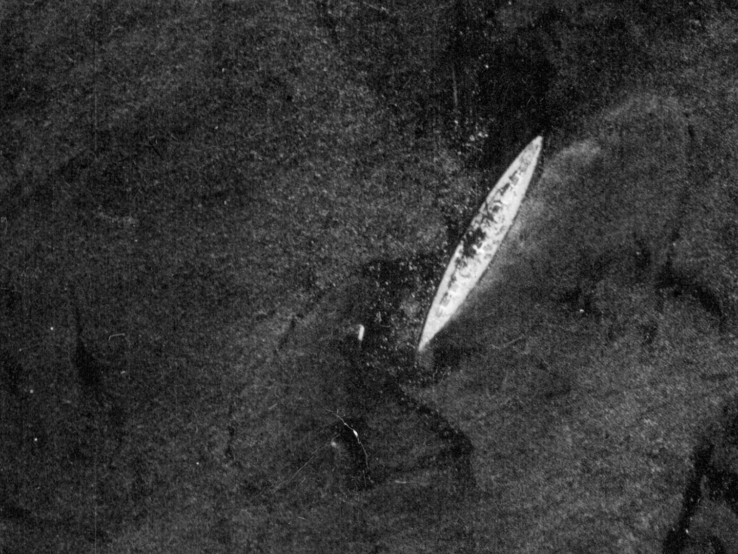 A historic black and white vertical aerial photo of the battleship Bismarck. The pale ship stands out on the dark sea.