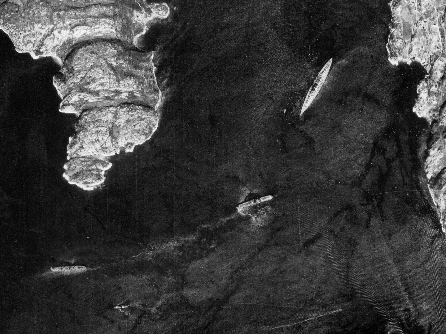 A black and white aerial photo of a Norwegian fjord with areas of rugged coastline visible at the right edge and top left corner. The largest ship – the Bismarck – is to the top right. 4 smaller ships can be seen towards the bottom of the image.