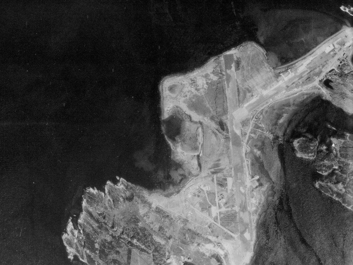 A historic black and white vertical aerial photo of a narrow, irregular island, with 2 runways running along spits of land close to the shore. The left side of the image is filled with the dark sea.