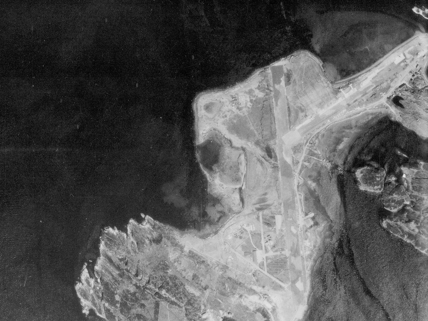 A historic black and white vertical aerial photo of a narrow, irregular island, with 2 runways running along spits of land close to the shore. The left side of the image is filled with the dark sea.