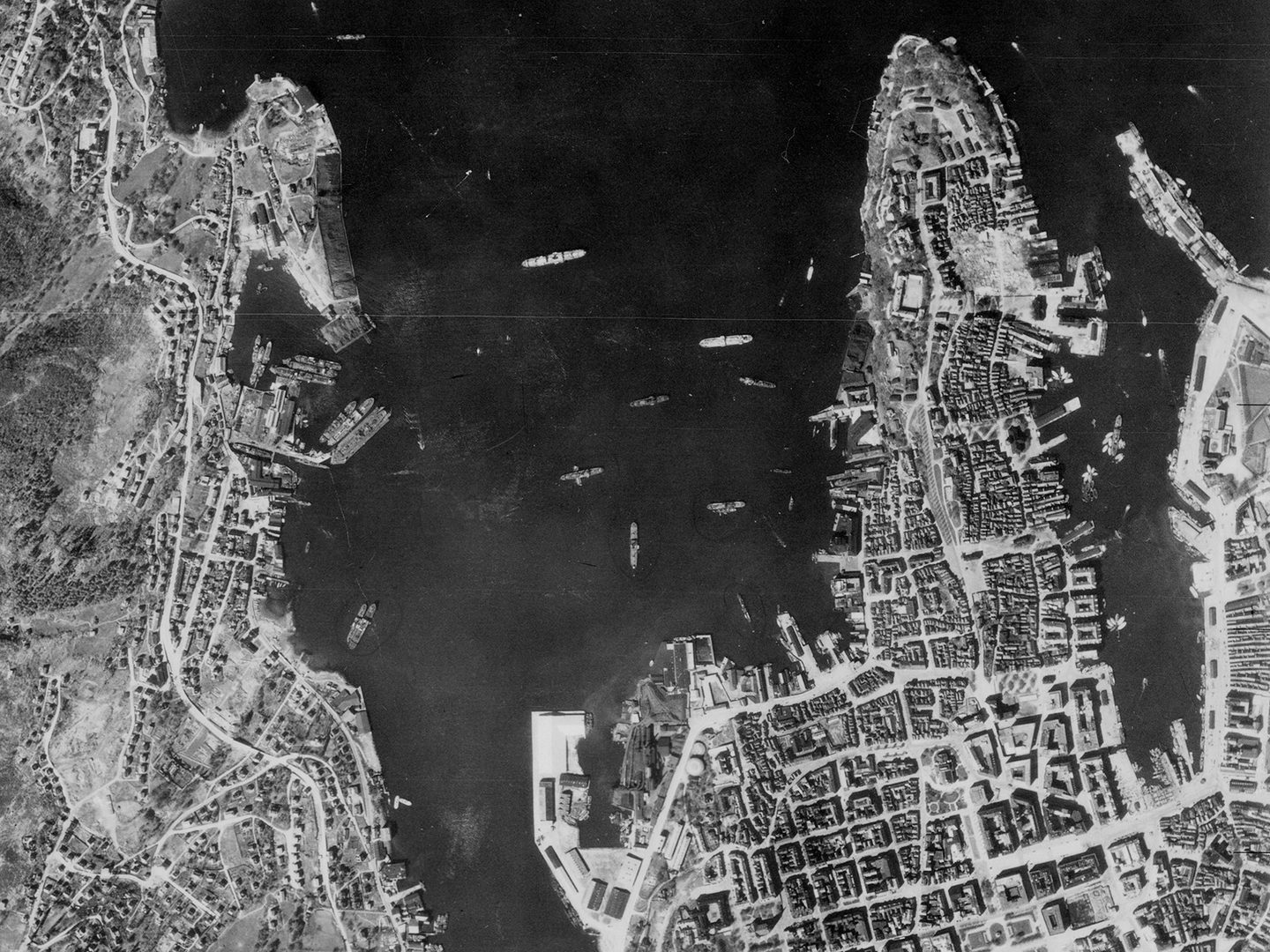 A historic black and white vertical aerial photo of a city built on 2 peninsulas running out into the sea. Between the fingers of land are numerous ships.