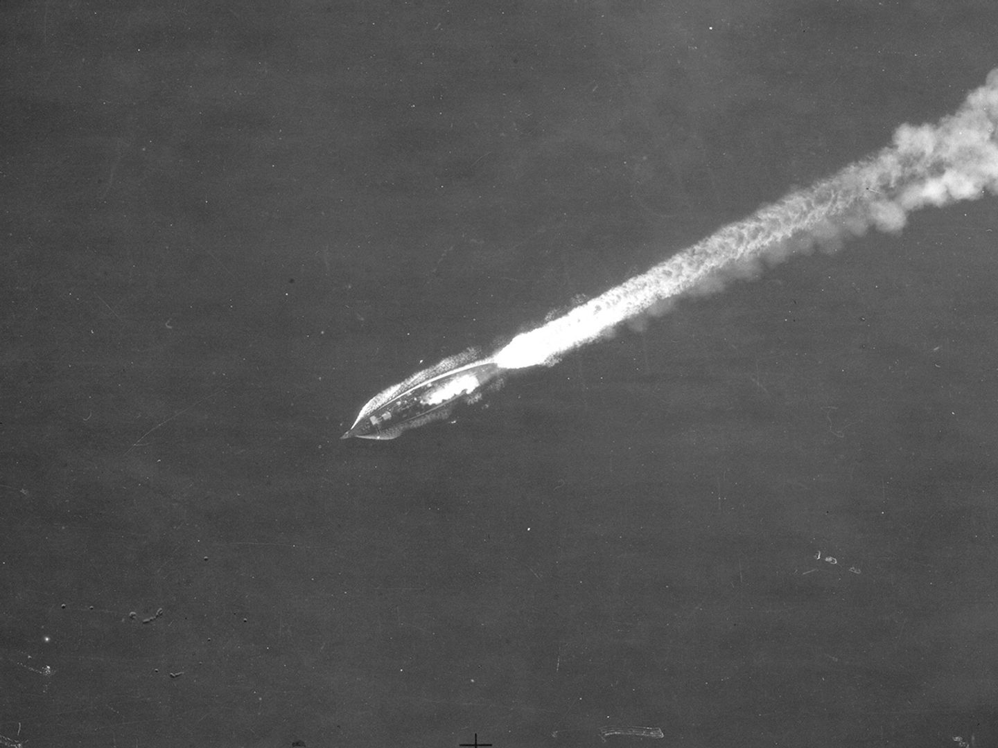A historic black and white vertical aerial photo of a battleship steaming across the sea from right to left. A white wake stretches out behind it towards the right.