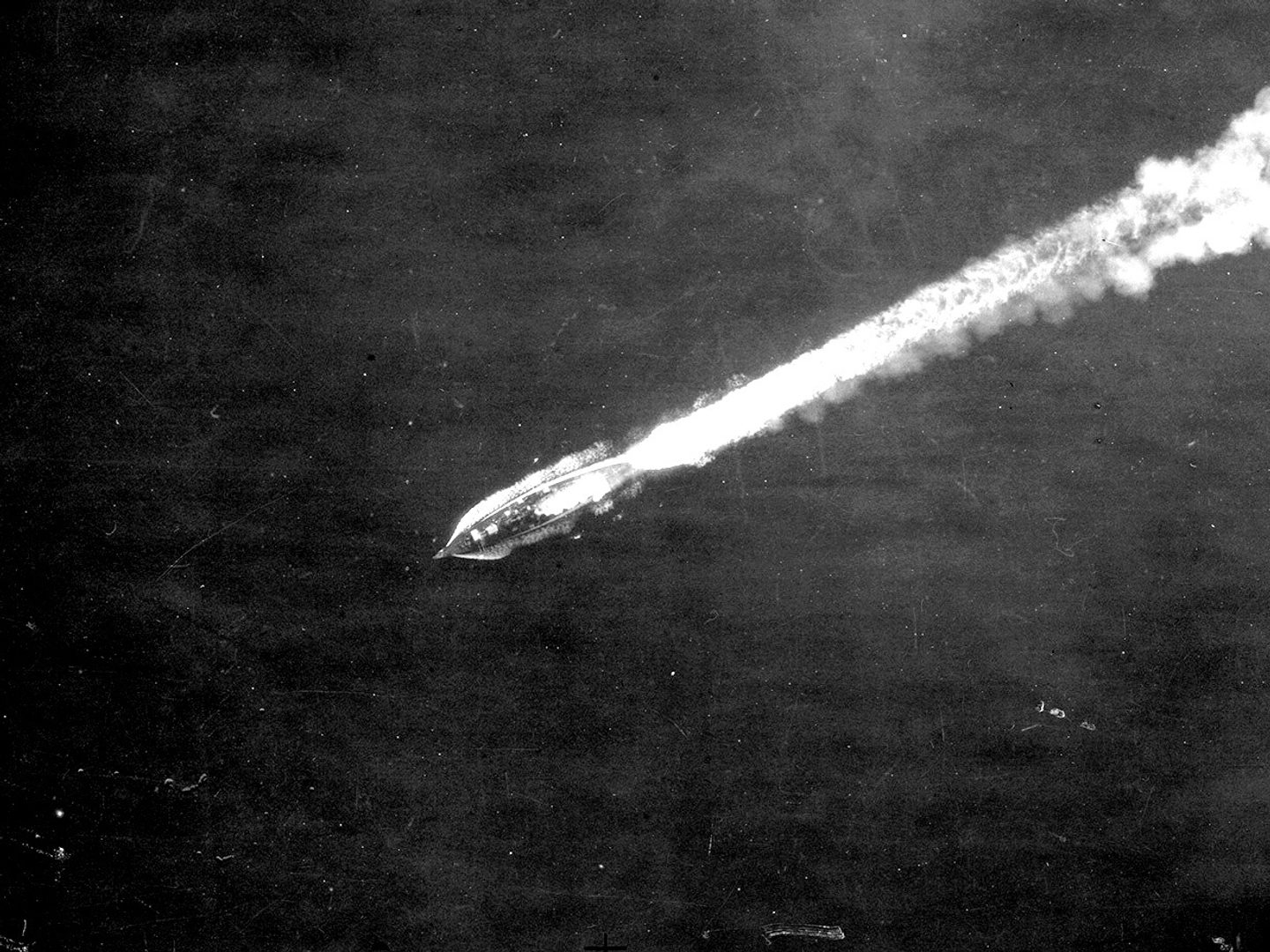 A historic black and white vertical aerial photo of a battleship steaming across the sea from right to left. A white wake stretches out behind it towards the right.