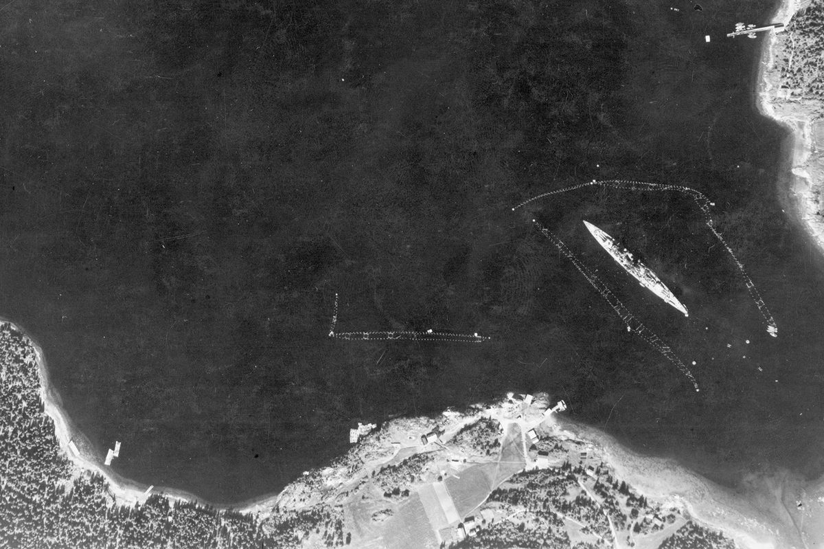A historic black and white vertical aerial photo of a large battleship moored, surrounded by torpedo netting. Areas of land are visible along the bottom edge and in the top right corner of the image.