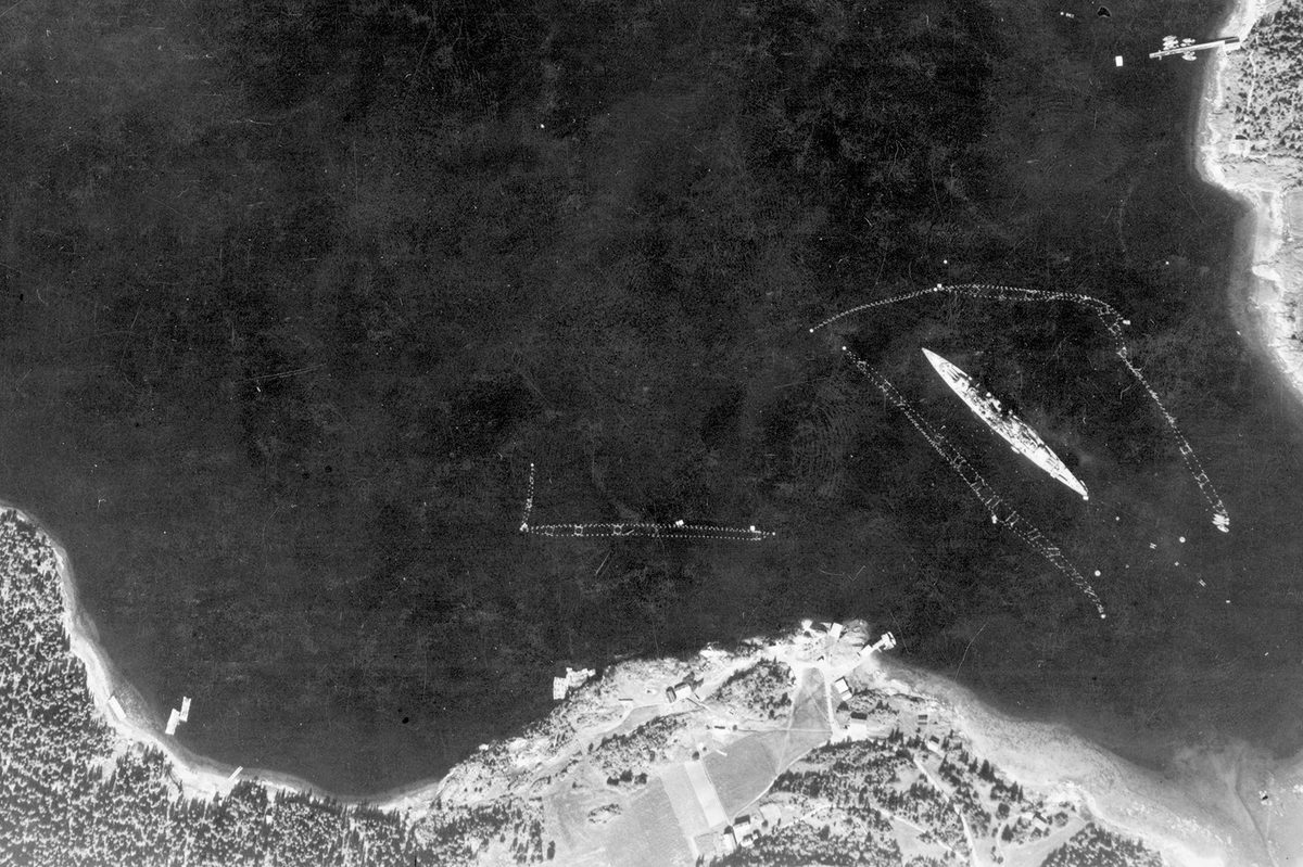 A historic black and white vertical aerial photo of a large battleship moored, surrounded by torpedo netting. Areas of land are visible along the bottom edge and in the top right corner of the image.