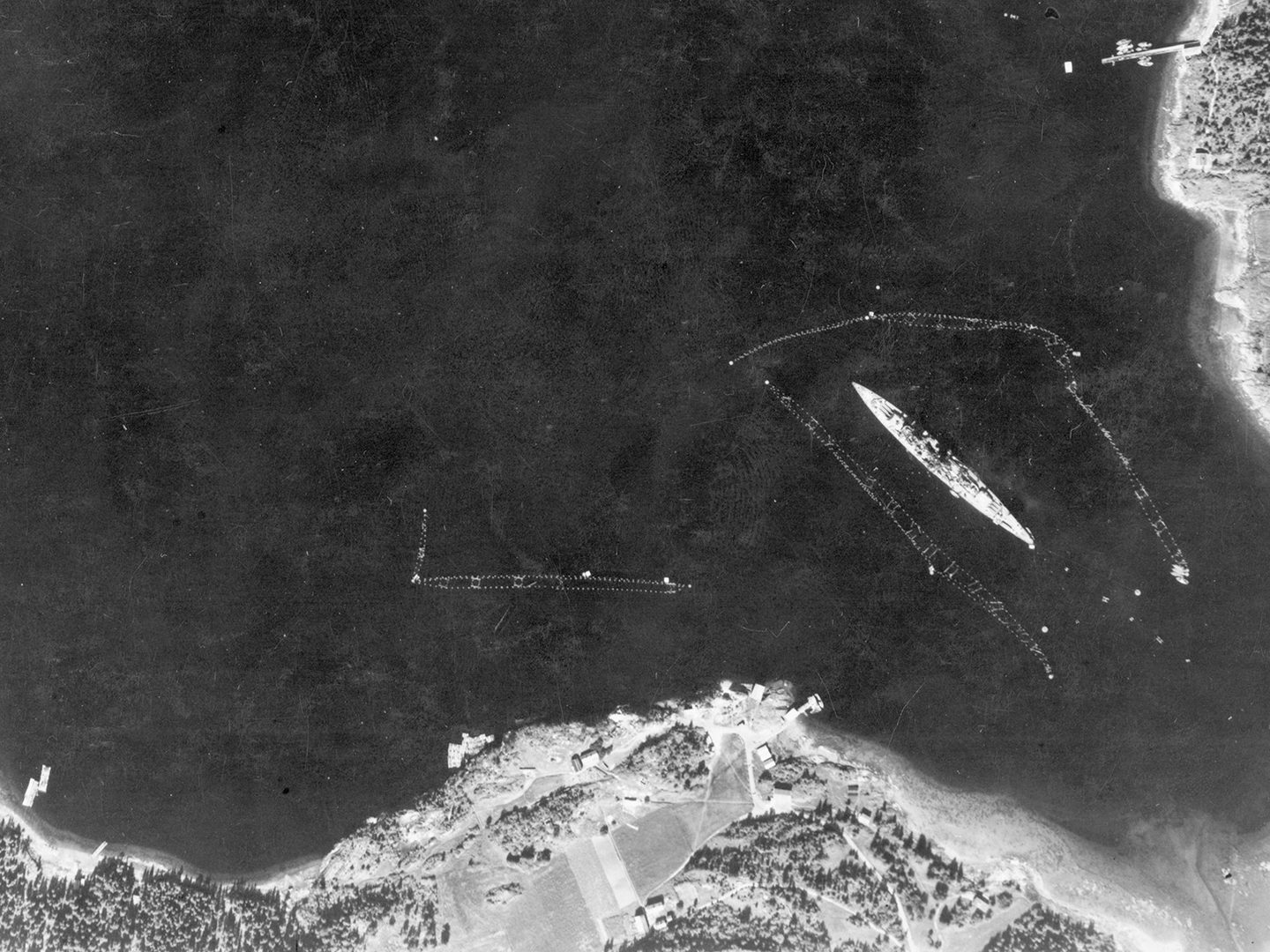 A historic black and white vertical aerial photo of a large battleship moored, surrounded by torpedo netting. Areas of land are visible along the bottom edge and in the top right corner of the image.