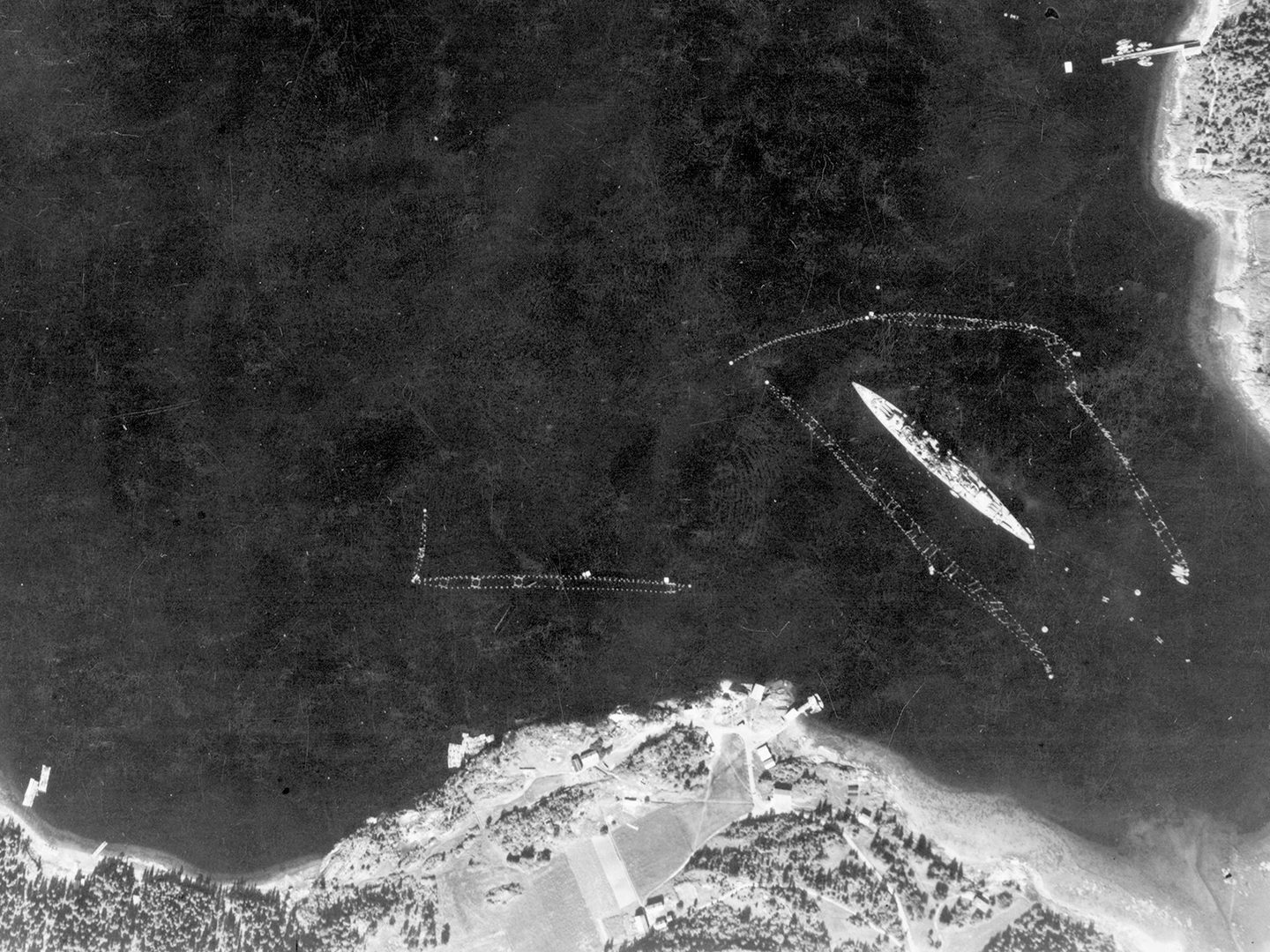 A historic black and white vertical aerial photo of a large battleship moored, surrounded by torpedo netting. Areas of land are visible along the bottom edge and in the top right corner of the image.