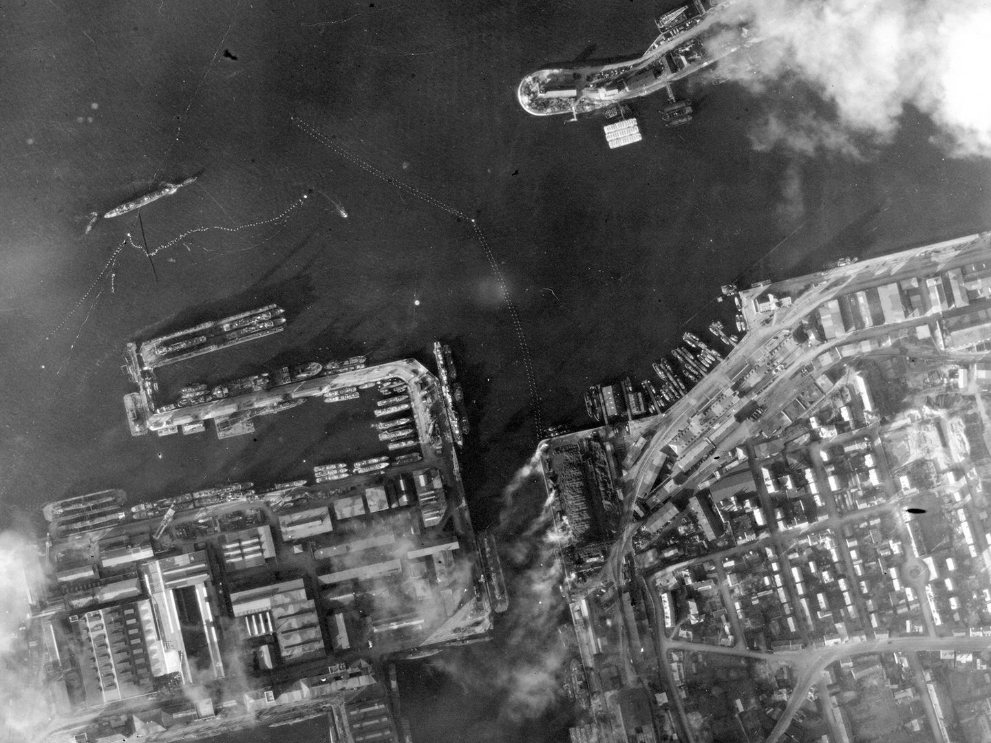 A historic black and white vertical aerial photo of a port. Docks lie along the lower edge of the image with vessels drawn up at them. Offshore is a larger ship surrounded by torpedo netting.
