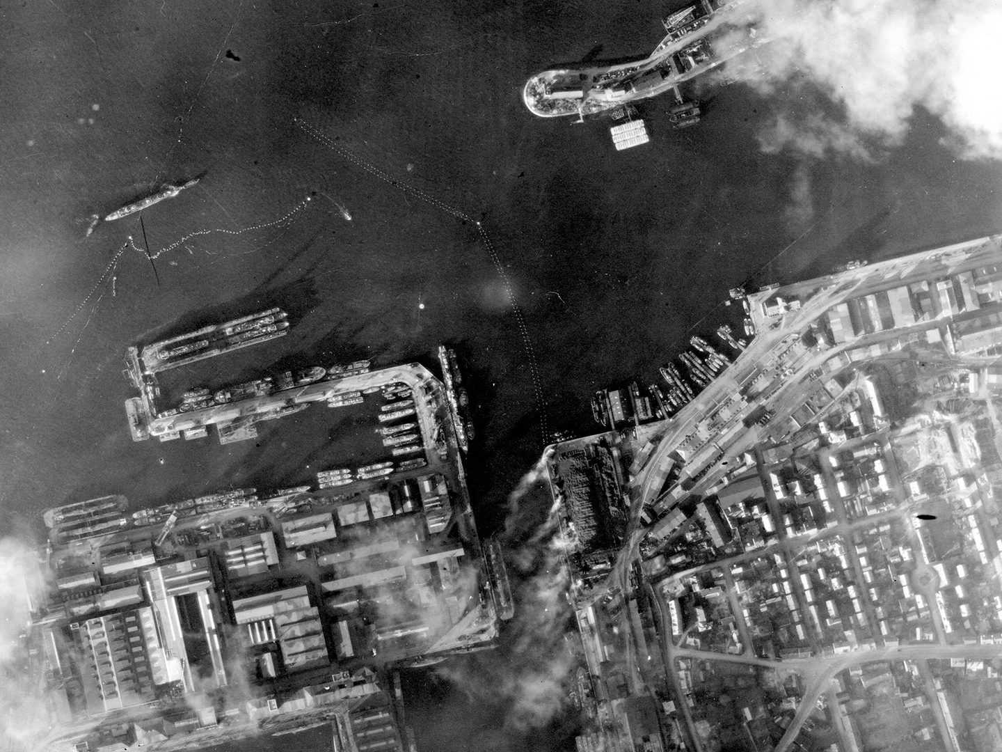 A historic black and white vertical aerial photo of a port. Docks lie along the lower edge of the image with vessels drawn up at them. Offshore is a larger ship surrounded by torpedo netting.