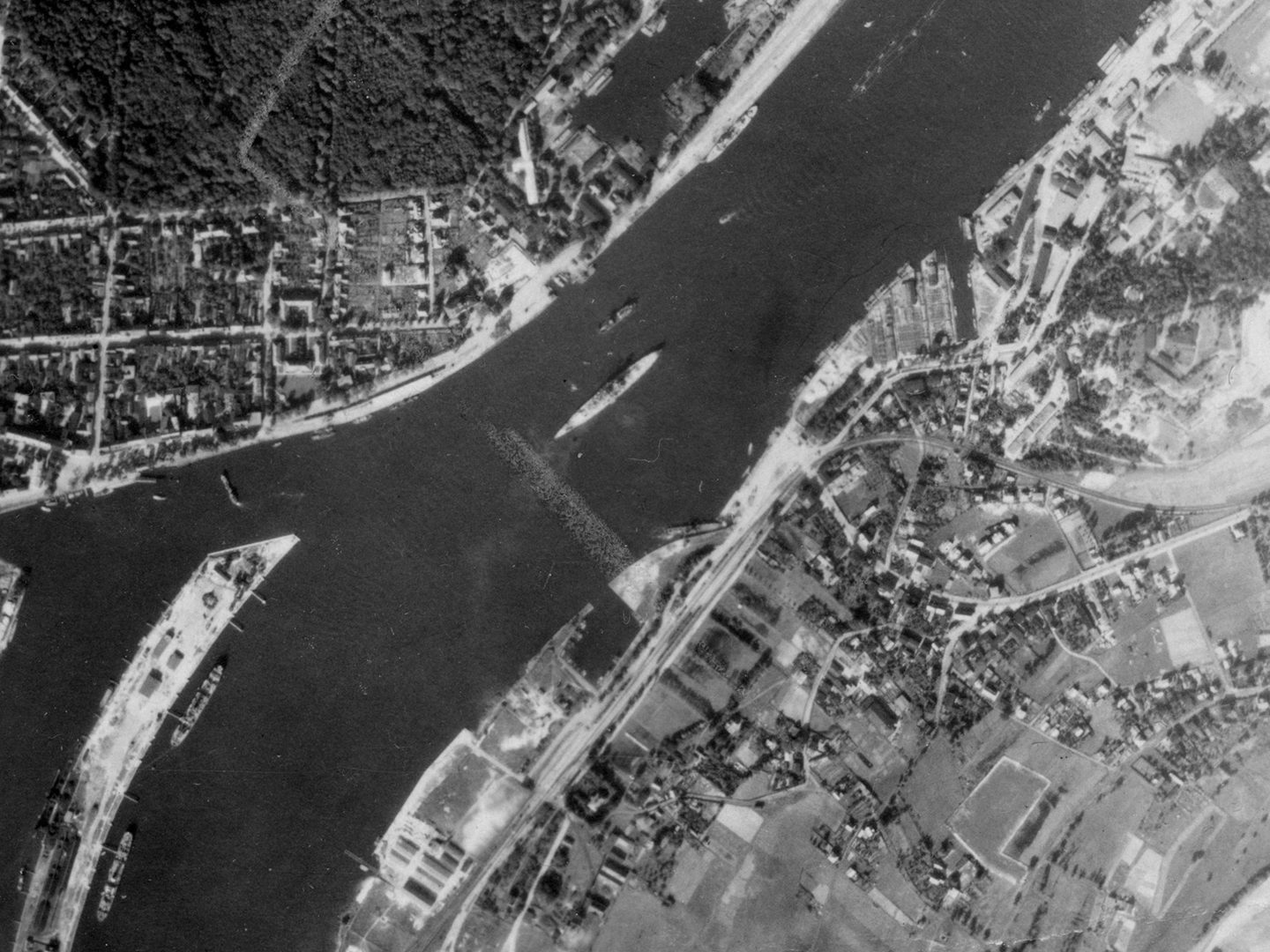 A historic black and white vertical aerial photo of a battleship in the middle of the river, running from top right to bottom left of the image. A mix of woods, fields and buildings cover the riverbanks.