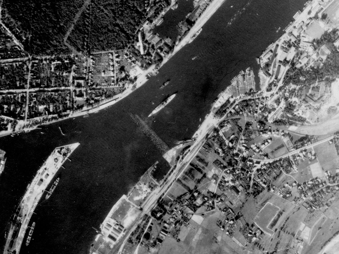 A historic black and white vertical aerial photo of a battleship in the middle of the river, running from top right to bottom left of the image. A mix of woods, fields and buildings cover the riverbanks.