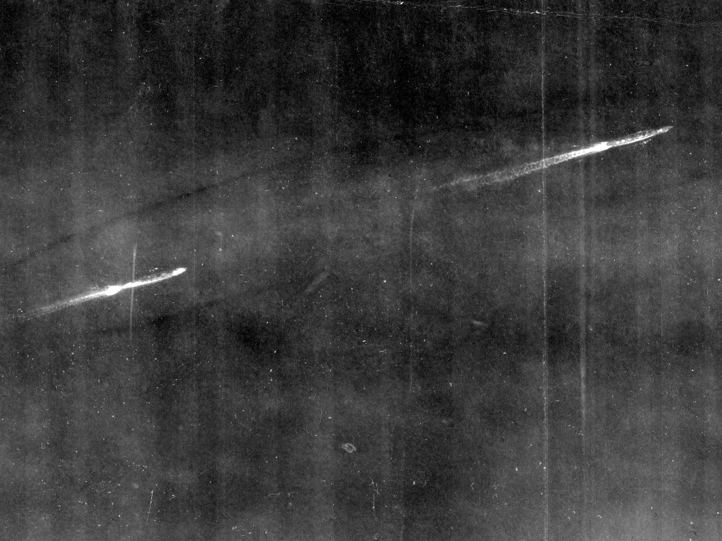 A historic black and white vertical aerial photo of two battleships steaming from left to right. Both have short white wakes stretching behind them.