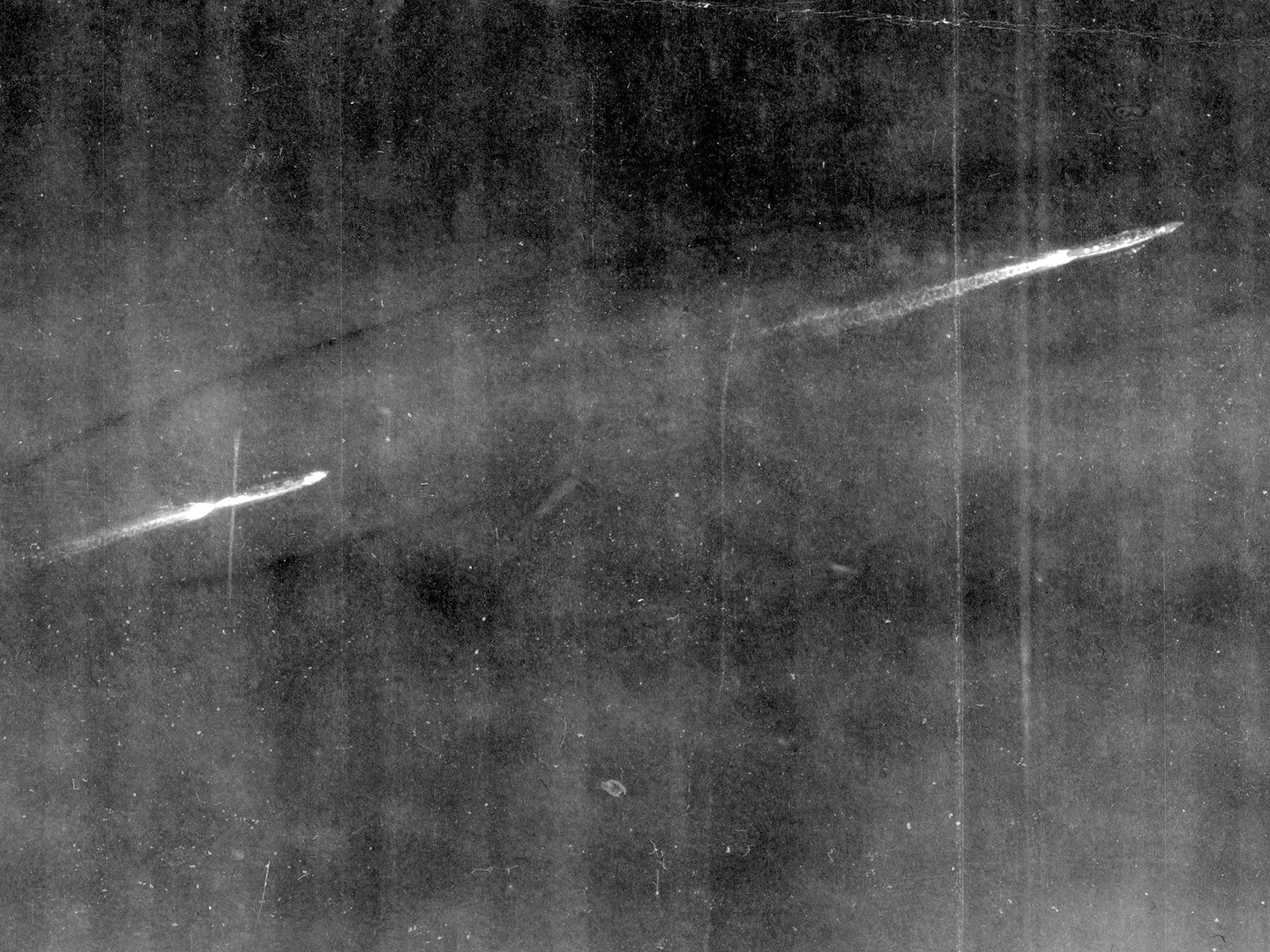 A historic black and white vertical aerial photo of two battleships steaming from left to right. Both have short white wakes stretching behind them.
