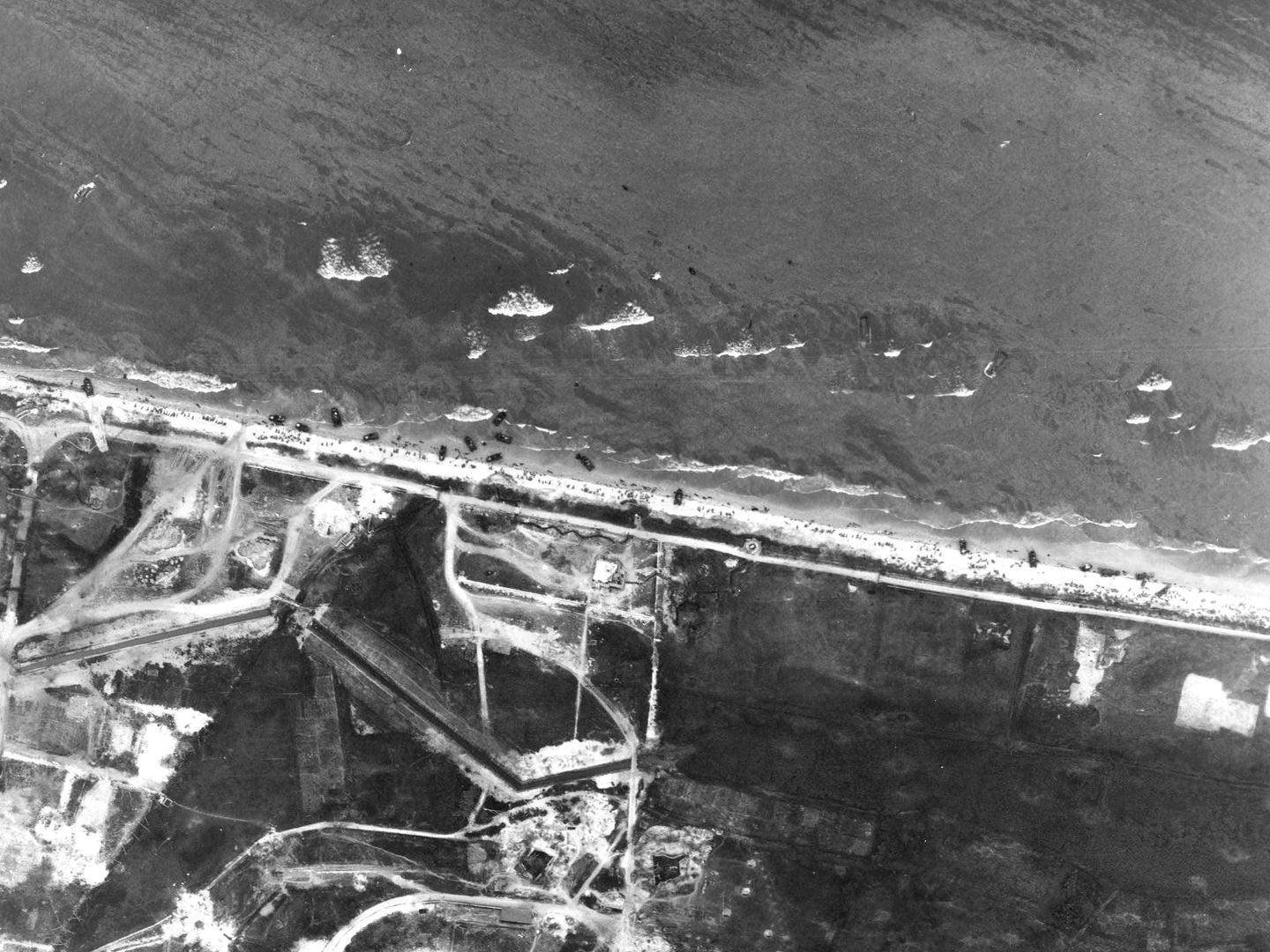 A black and white aerial photo of numerous small landing craft pulled up on a beach covered in people. Waves are washing in from the sea. The land beyond the beach at the bottom of the image looks scarred from construction work and shelling/bombing.