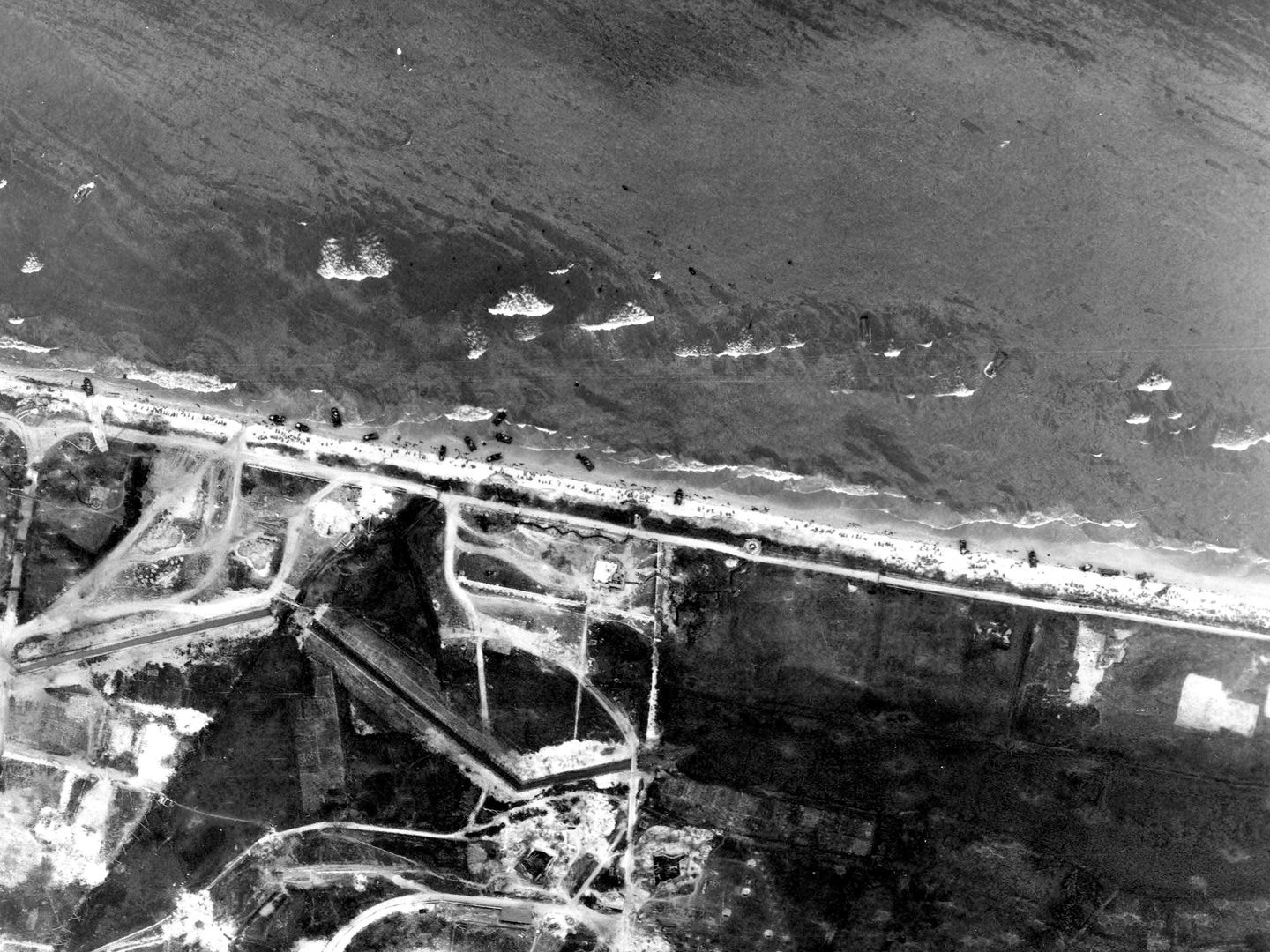 A black and white aerial photo of numerous small landing craft pulled up on a beach covered in people. Waves are washing in from the sea. The land beyond the beach at the bottom of the image looks scarred from construction work and shelling/bombing.