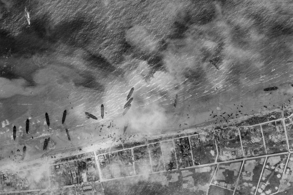 A black and white aerial photo of about a dozen ships pulled up on a beach. The land beyond the beach has been divided up into a grid, and the scene is slightly obscured by clouds or smoke.