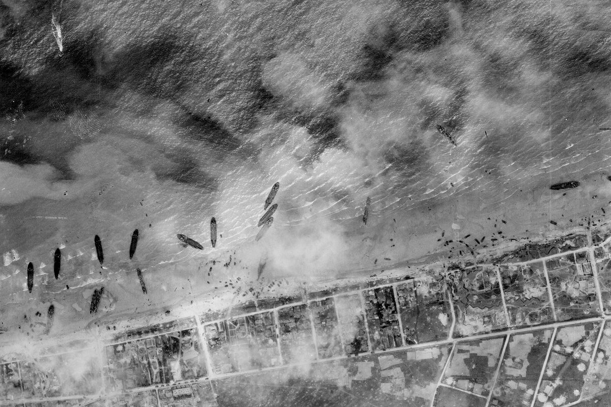 A black and white aerial photo of about a dozen ships pulled up on a beach. The land beyond the beach has been divided up into a grid, and the scene is slightly obscured by clouds or smoke.