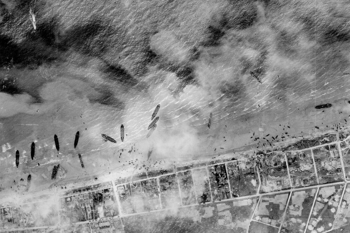 A historic black and white vertical aerial photo of about a dozen ships pulled up on a beach. The land beyond the beach has been divided up into a grid, and the scene is slightly obscured by clouds or smoke.