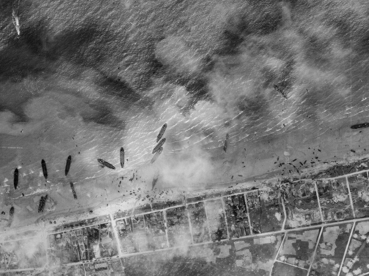 A black and white aerial photo of about a dozen ships pulled up on a beach. The land beyond the beach has been divided up into a grid, and the scene is slightly obscured by clouds or smoke.