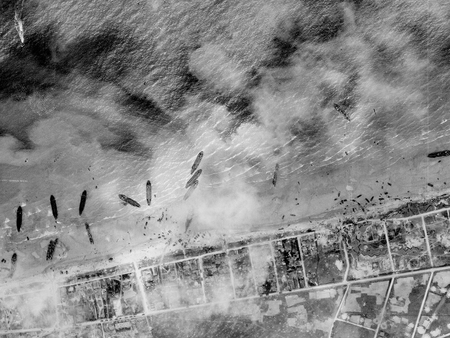 A historic black and white vertical aerial photo of about a dozen ships pulled up on a beach. The land beyond the beach has been divided up into a grid, and the scene is slightly obscured by clouds or smoke.
