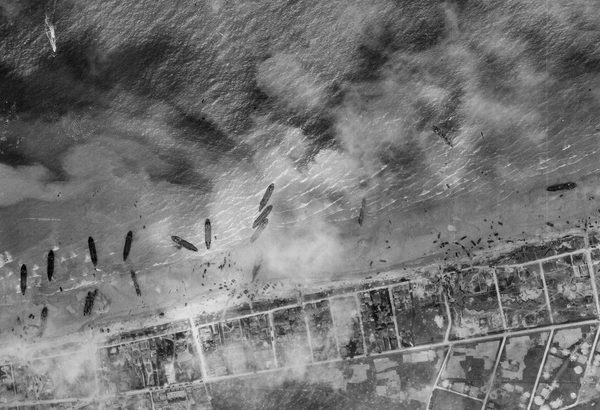 A black and white aerial photo of about a dozen ships pulled up on a beach. The land beyond the beach has been divided up into a grid, and the scene is slightly obscured by clouds or smoke.