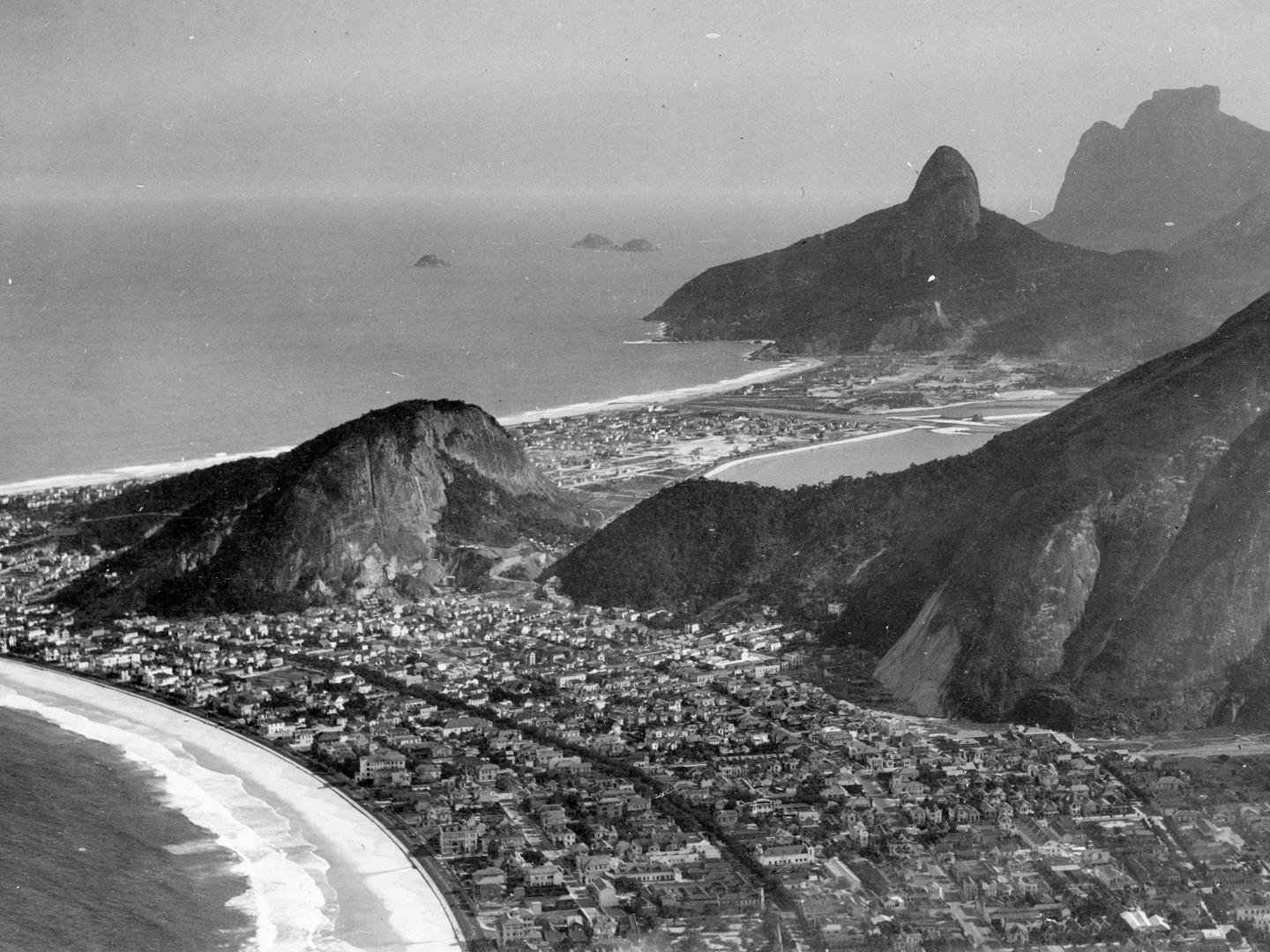 A black and white oblique aerial photo of a city spread along a sandy shoreline. Waves crash on the shore. The city buildings appear mostly as white low-rises. Several forest-covered rocky domes rise above the city.