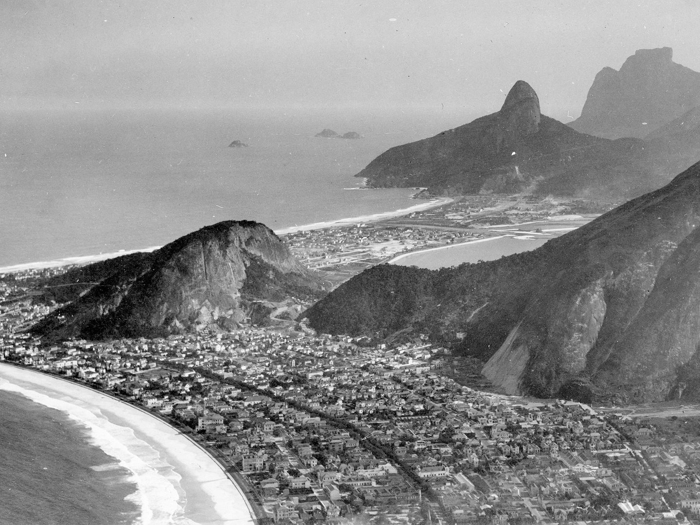 A black and white oblique aerial photo of a city spread along a sandy shoreline. Waves crash on the shore. The city buildings appear mostly as white low-rises. Several forest-covered rocky domes rise above the city.