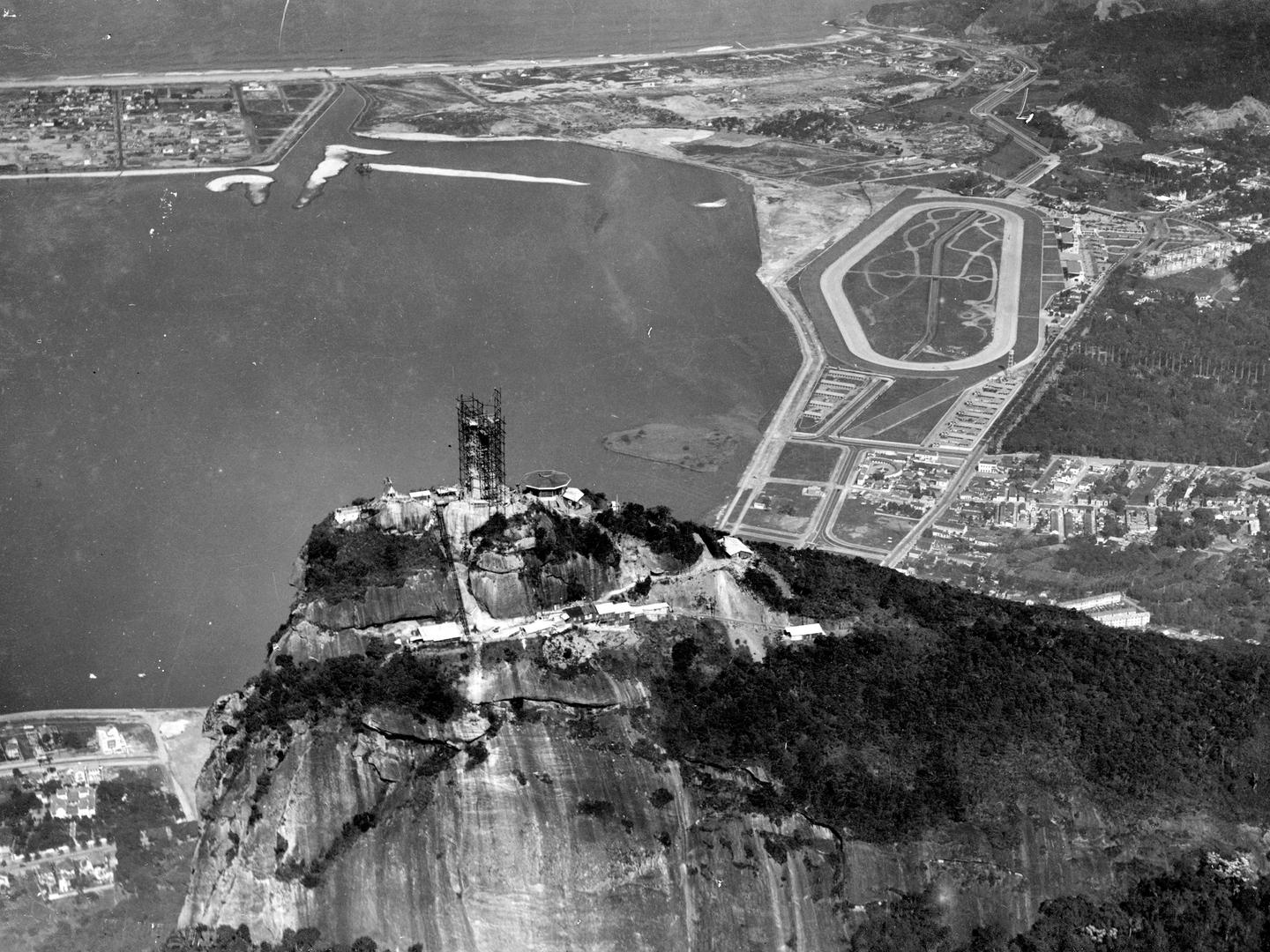 A black and white oblique aerial photo of a rocky mountain peak. At its summit a huge statue is swathed in scaffolding. Beyond the statue is a large bay. Scattered areas of buildings are visible on the surrounding shores.