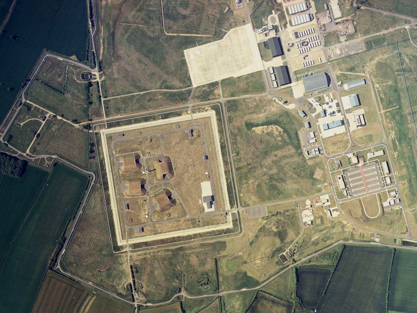 A colour aerial photo of the green fields of an airfield complex. To the left are 4 grass-covered missile bunkers surrounded by a square enclosure. To the right are huts and buildings in a variety of sizes. The different areas are linked by roads.