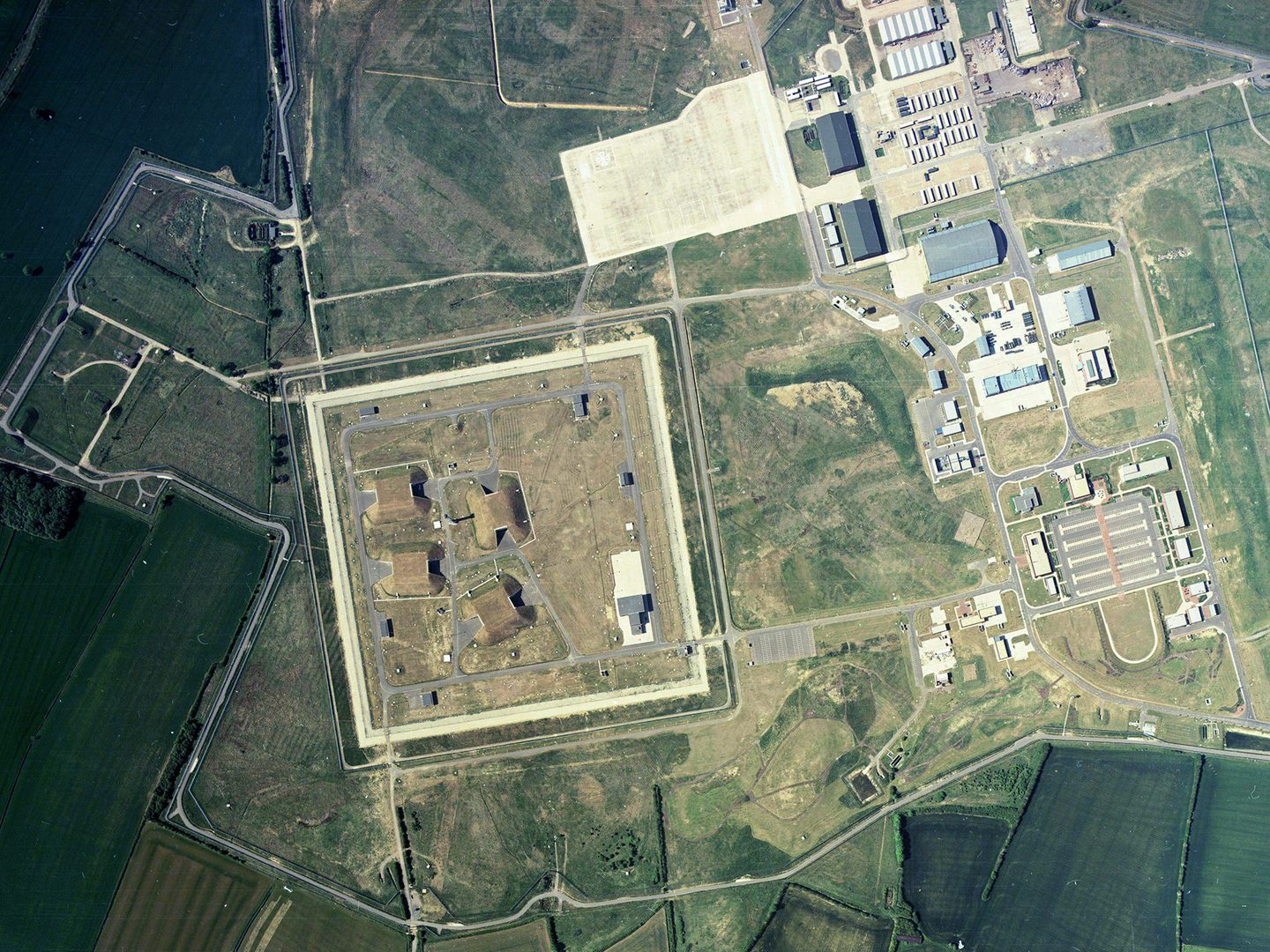 A colour aerial photo of the green fields of an airfield complex. To the left are 4 grass-covered missile bunkers surrounded by a square enclosure. To the right are huts and buildings in a variety of sizes. The different areas are linked by roads.