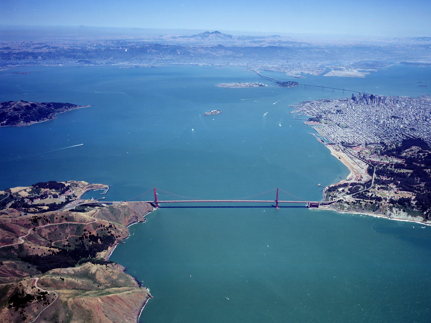 A colour aerial photo of a red suspension bridge joining two areas of land across a large bay. A large city is visible on the right. The sea stretches into the distance, where on the horizon forested, mountainous land is visible.
