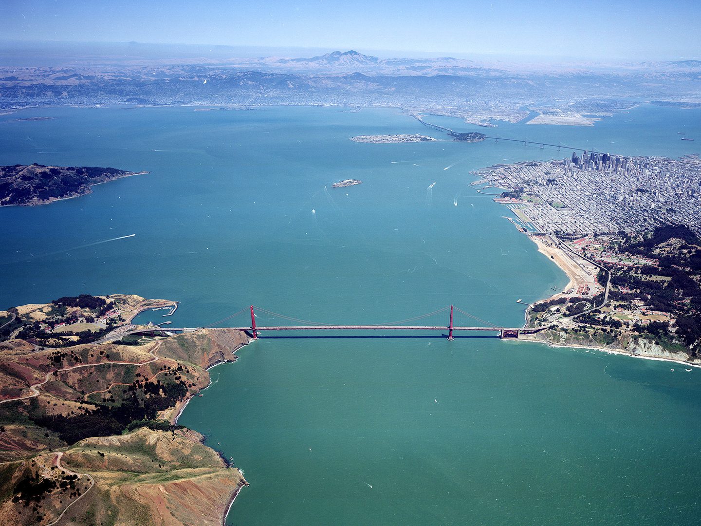 A colour aerial photo of a red suspension bridge joining two areas of land across a large bay. A large city is visible on the right. The sea stretches into the distance, where on the horizon forested, mountainous land is visible.