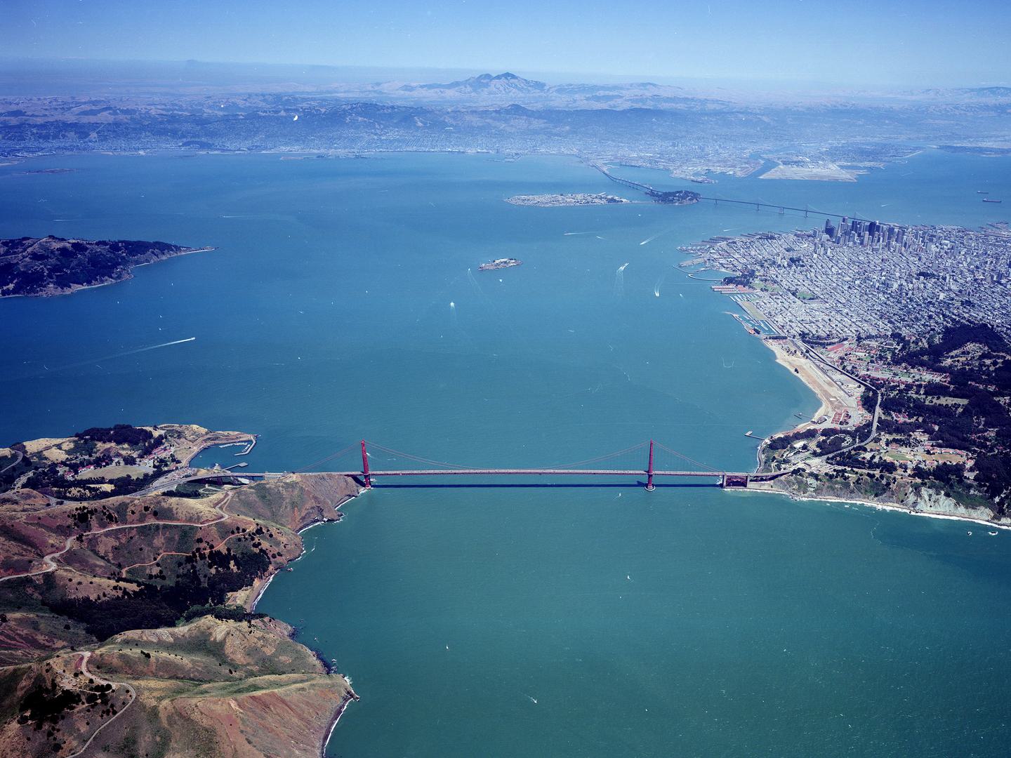 A colour aerial photo of a red suspension bridge joining two areas of land across a large bay. A large city is visible on the right. The sea stretches into the distance, where on the horizon forested, mountainous land is visible.
