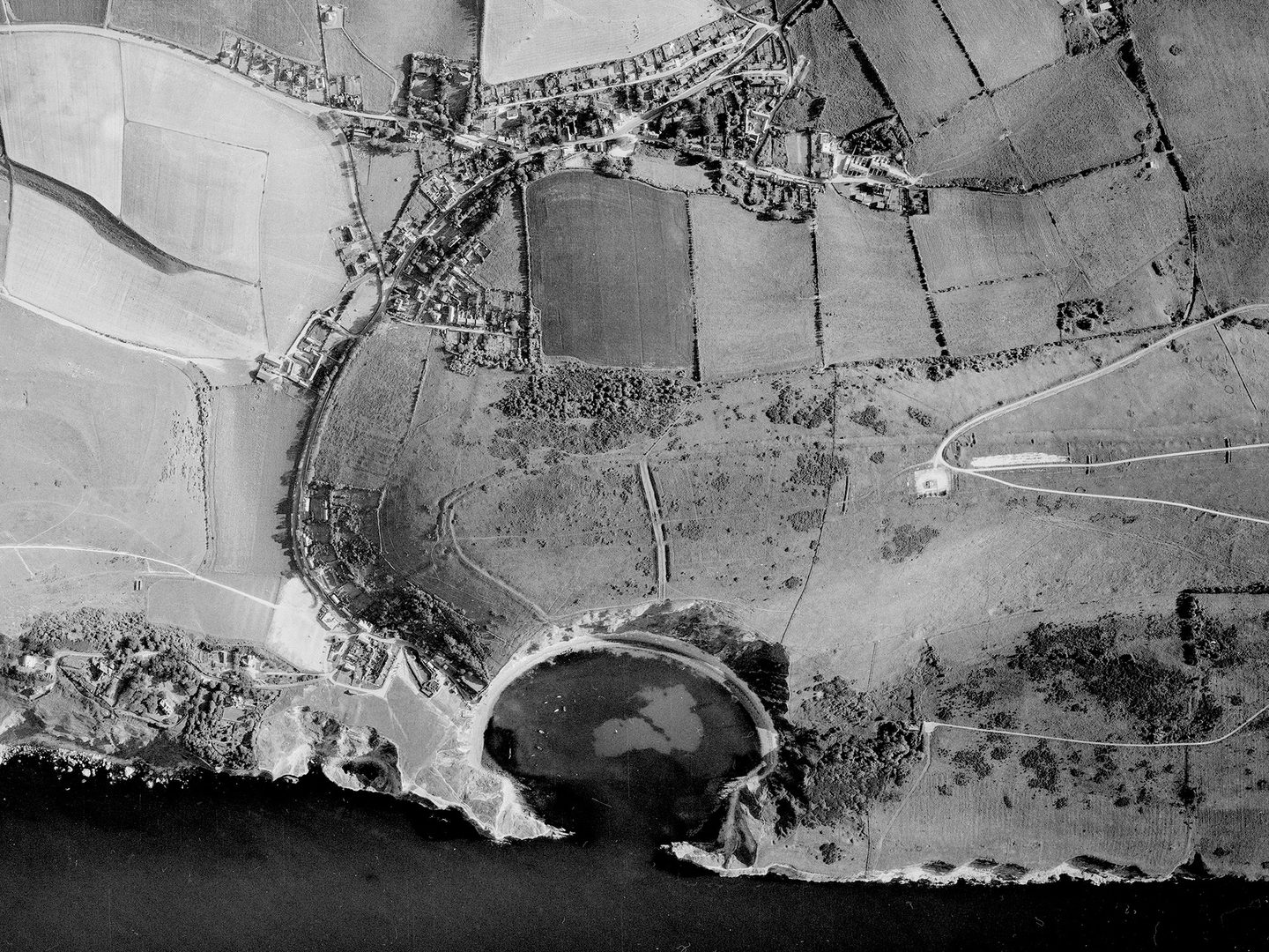 A black and white aerial photo of a coastline. The coast is formed of white cliffs which indent to create a dramatic semi-circular bay. Beyond the bay are fields. A road leads from the bay through the fields to a small village.