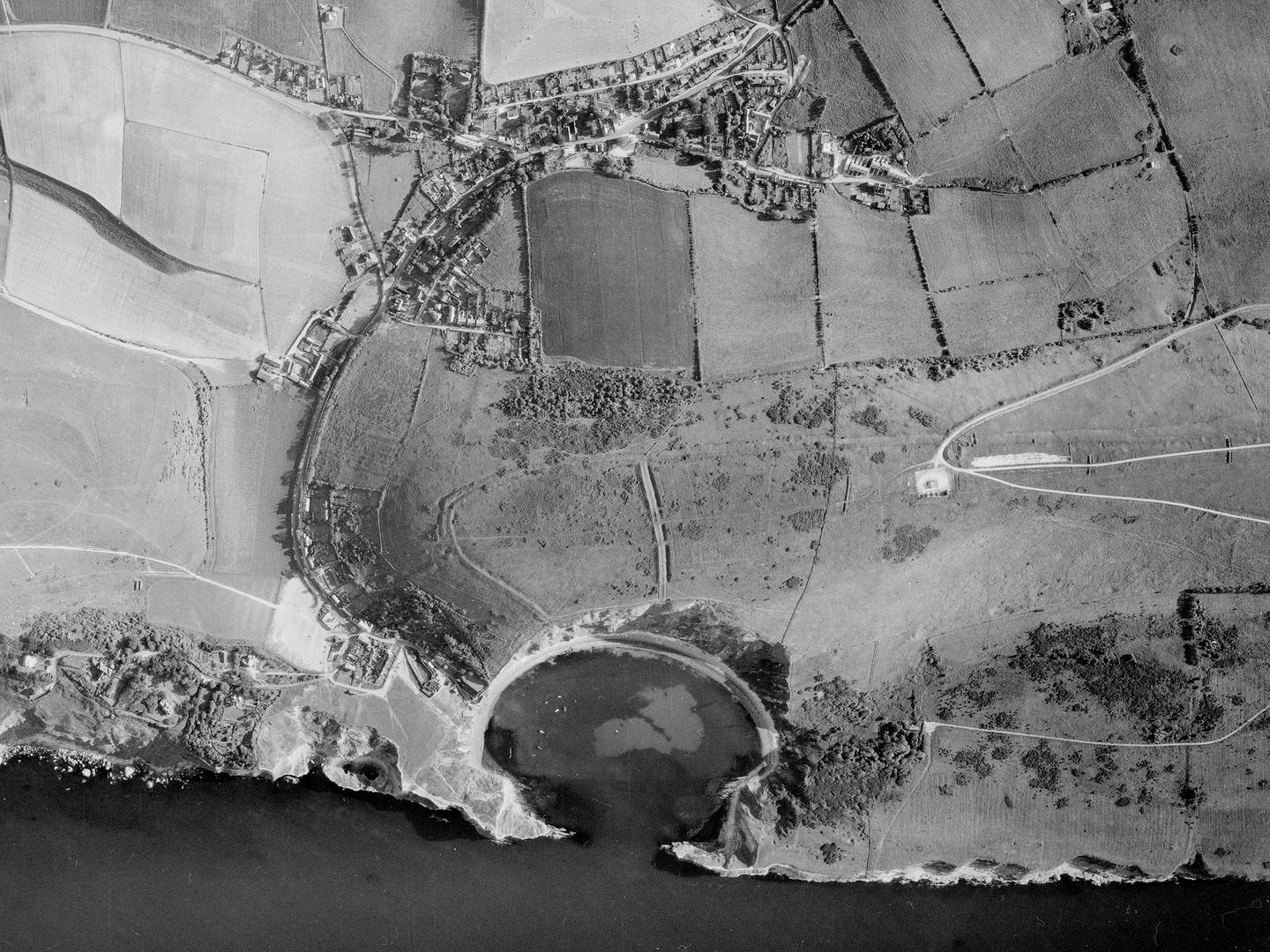 A black and white aerial photo of a coastline. The coast is formed of white cliffs which indent to create a dramatic semi-circular bay. Beyond the bay are fields. A road leads from the bay through the fields to a small village.
