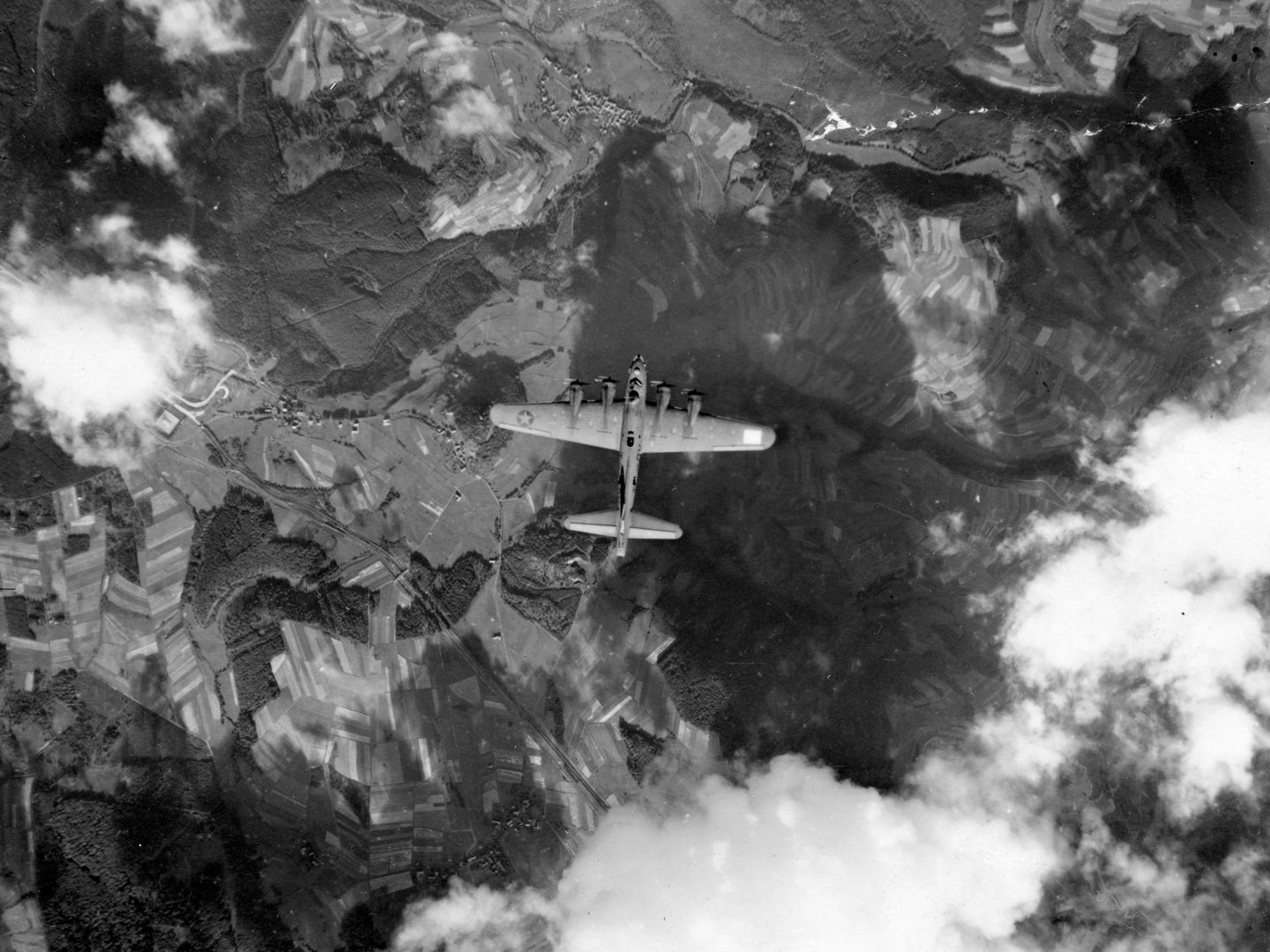 A historic black and white vertical aerial photo of a 4-winged bomber flying over a wooded landscape. A few white clouds are visible around the aircraft.