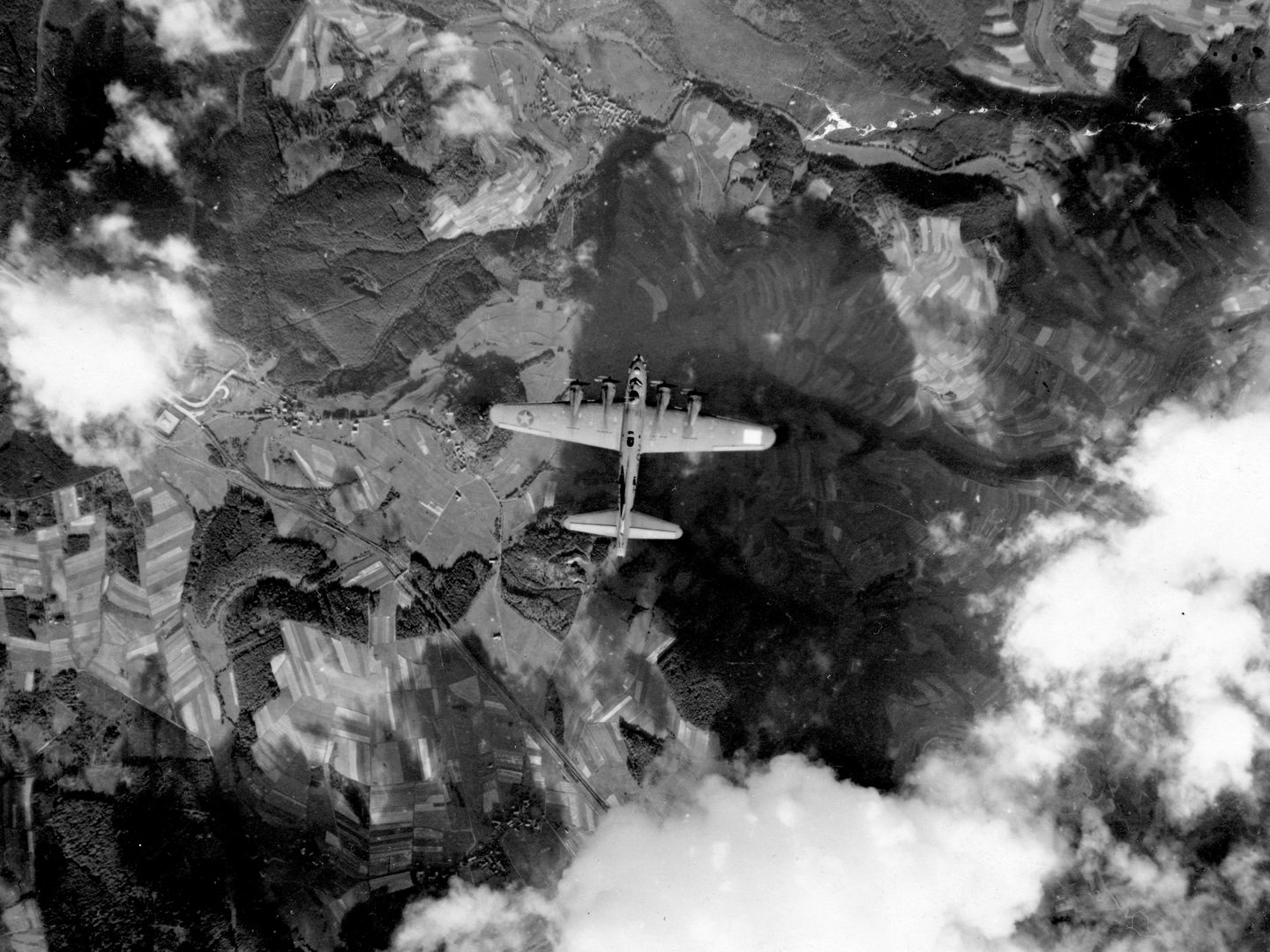 A historic black and white vertical aerial photo of a 4-winged bomber flying over a wooded landscape. A few white clouds are visible around the aircraft.
