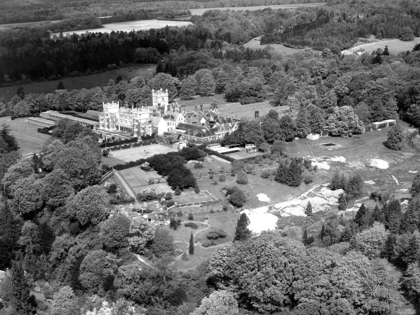 A historic black and white oblique aerial photo of a grand white stately home in mock Tudor style. It is surrounded by extensive grounds which contain numerous huts and other temporary buildings. The landscape around the grounds is heavily wooded.