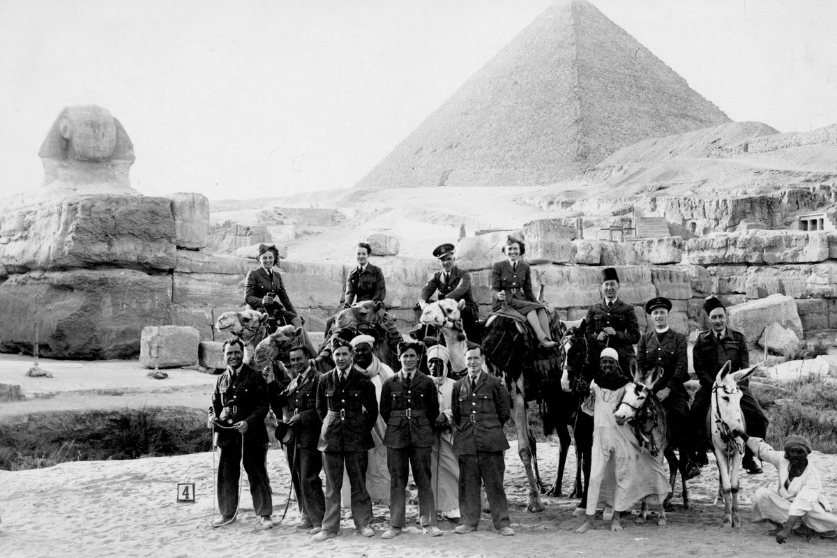 A historic black and white photo of a group of people in front of the Great Sphinx and Great Pyramid of Giza. Most of the group are European military personnel (men and women) in uniform, some standing, some sitting on camels and some sitting on donkeys. Several Egyptian men in white robes are also present.
