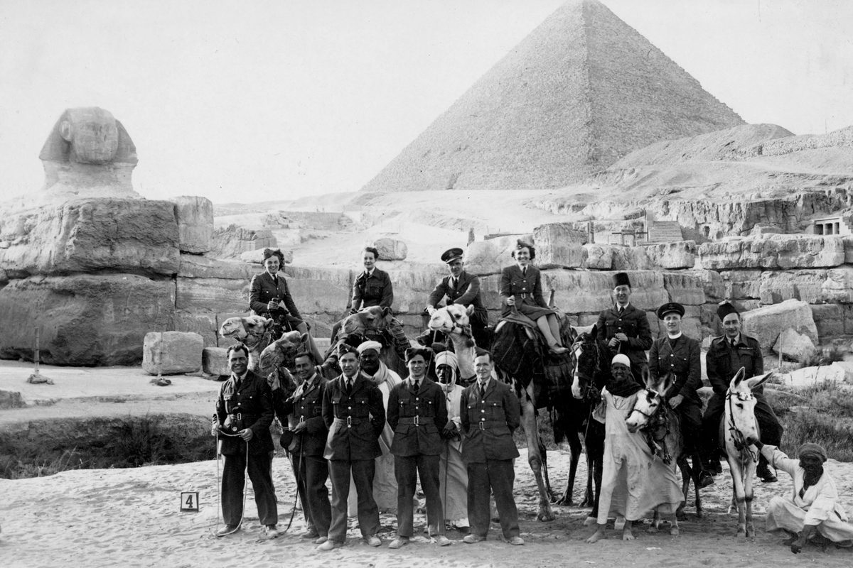 A historic black and white photo of a group of people in front of the Great Sphinx and Great Pyramid of Giza. Most of the group are European military personnel (men and women) in uniform, some standing, some sitting on camels and some sitting on donkeys. Several Egyptian men in white robes are also present.