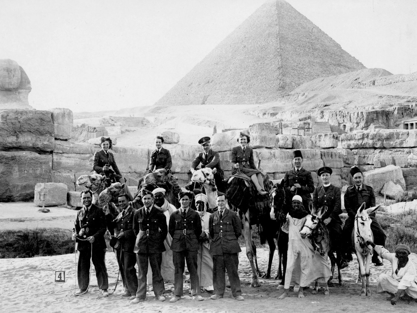 A historic black and white photo of a group of people in front of the Great Sphinx and Great Pyramid of Giza. Most of the group are European military personnel (men and women) in uniform, some standing, some sitting on camels and some sitting on donkeys. Several Egyptian men in white robes are also present.