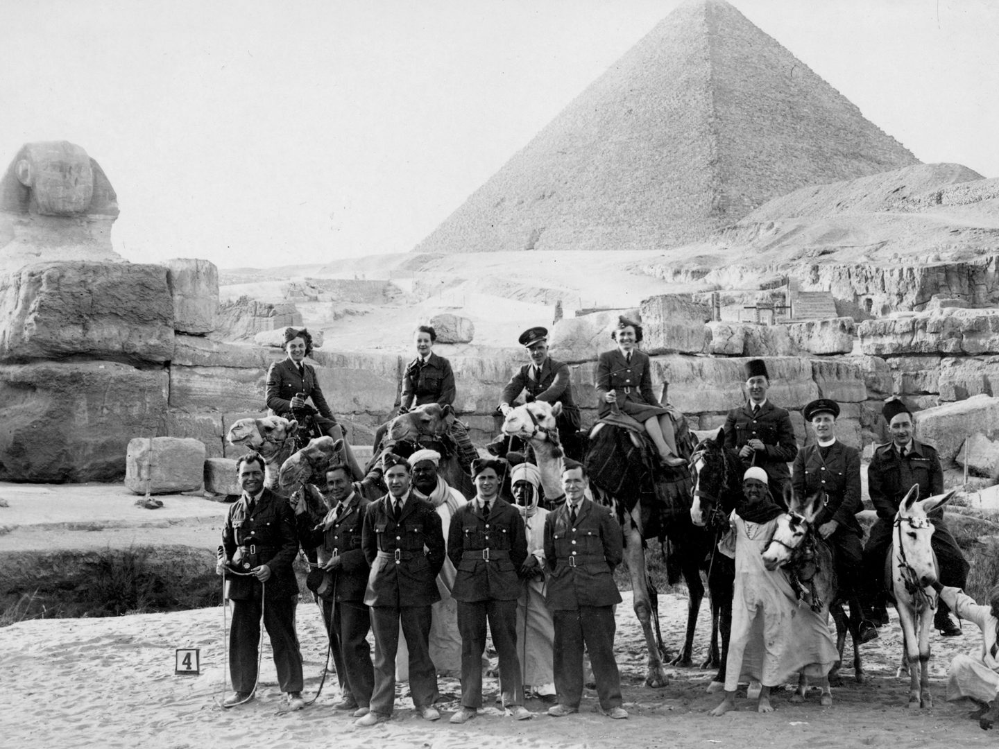 A historic black and white photo of a group of people in front of the Great Sphinx and Great Pyramid of Giza. Most of the group are European military personnel (men and women) in uniform, some standing, some sitting on camels and some sitting on donkeys. Several Egyptian men in white robes are also present.