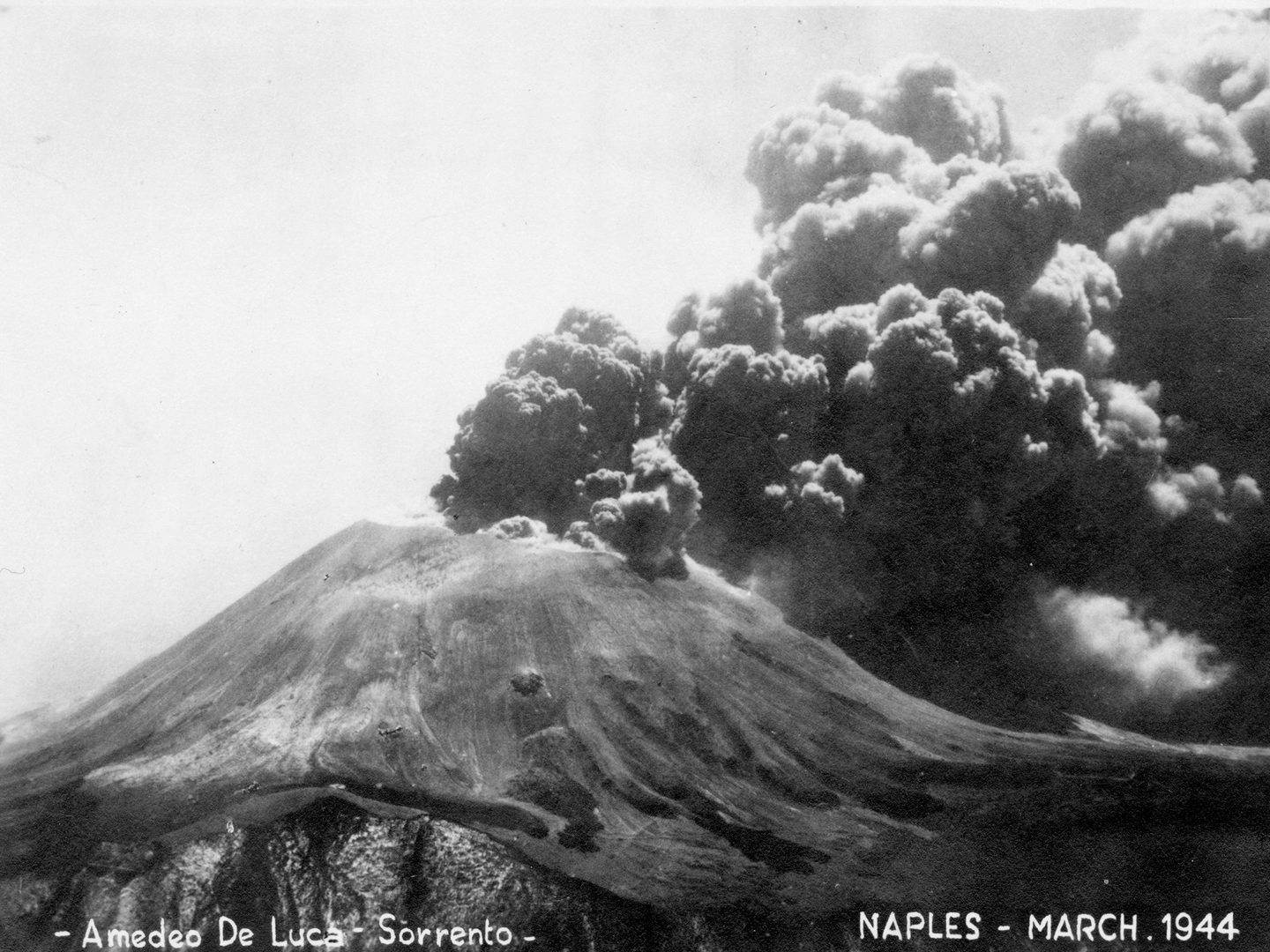 A historic black and white postcard of Mount Vesuvius erupting. Clouds of black ash are rising from its summit. Text along the bottom of the postcard specifies the photographer (Amedeo de Luca of Sorrento), the location (Naples) and the date (March 1944).