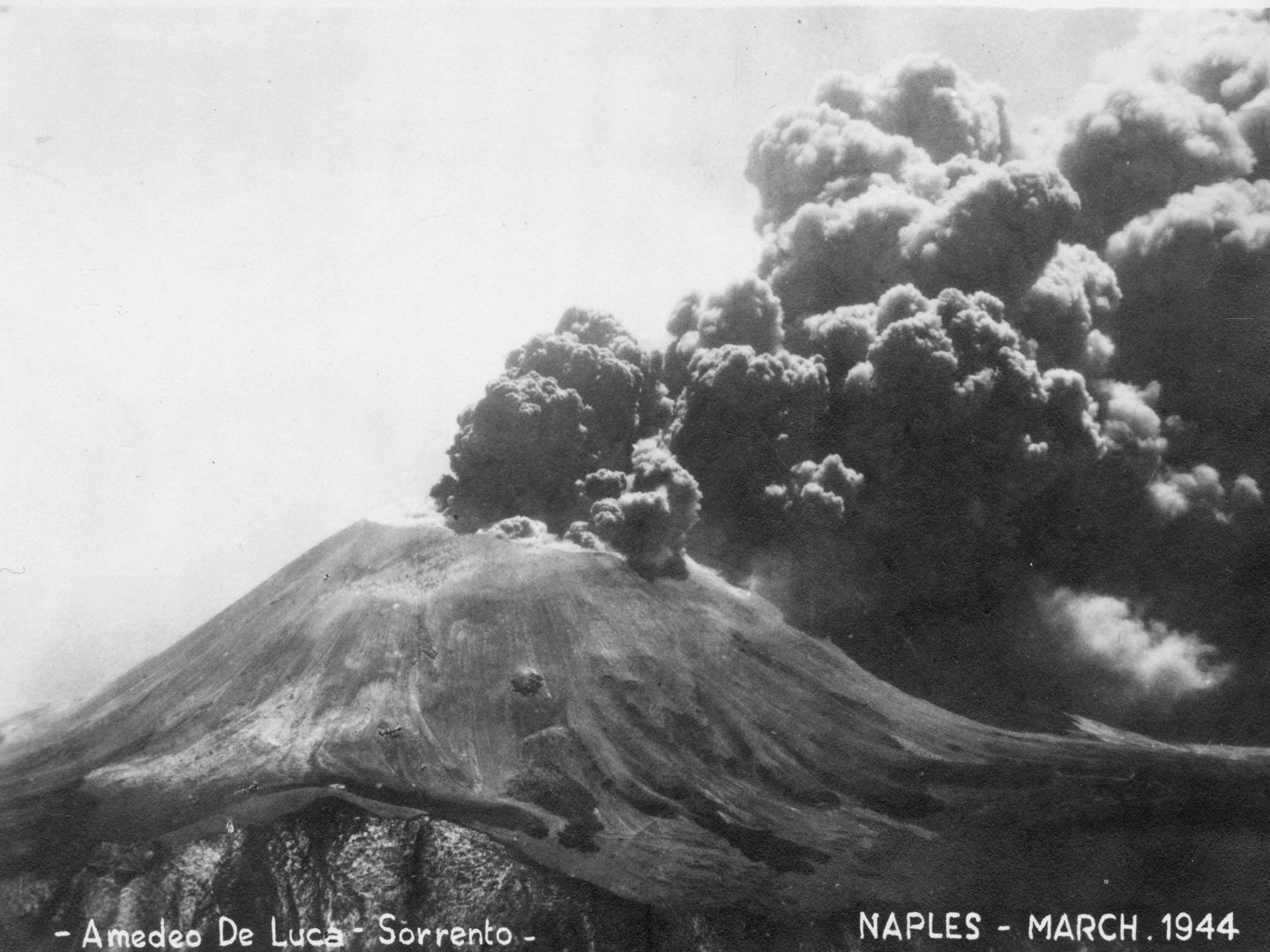 A historic black and white postcard of Mount Vesuvius erupting. Clouds of black ash are rising from its summit. Text along the bottom of the postcard specifies the photographer (Amedeo de Luca of Sorrento), the location (Naples) and the date (March 1944).