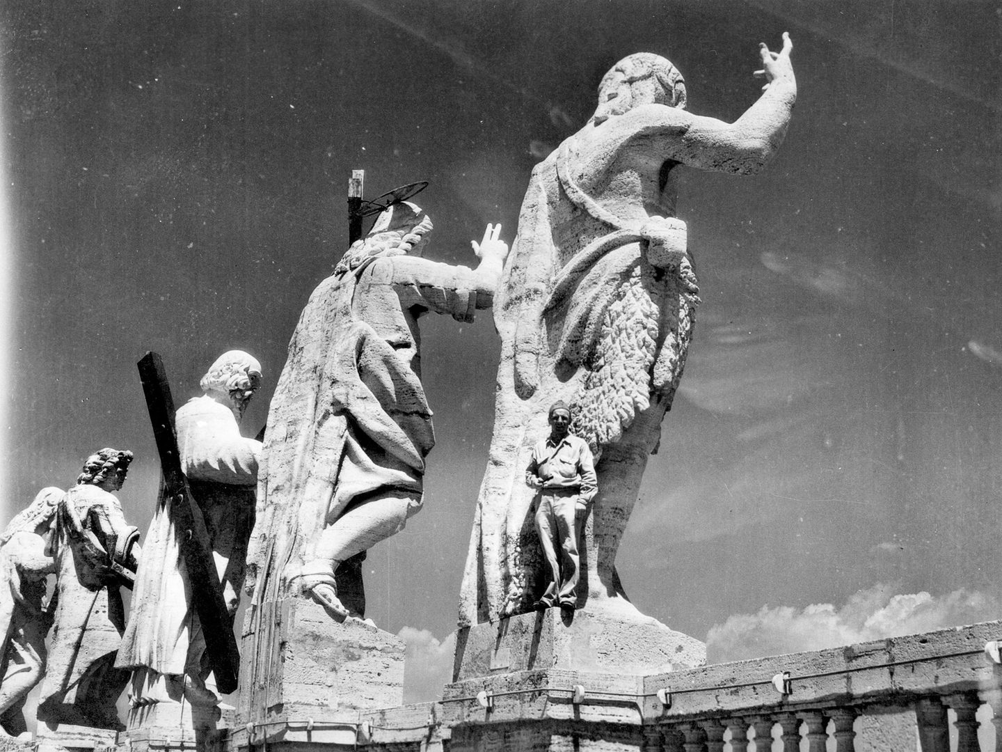 A historic black and white photo of 4 enormous sculptures seen from behind. On the plinth of the nearest sculpture stands Donald Beer, dwarfed by the scale of the figure above him.