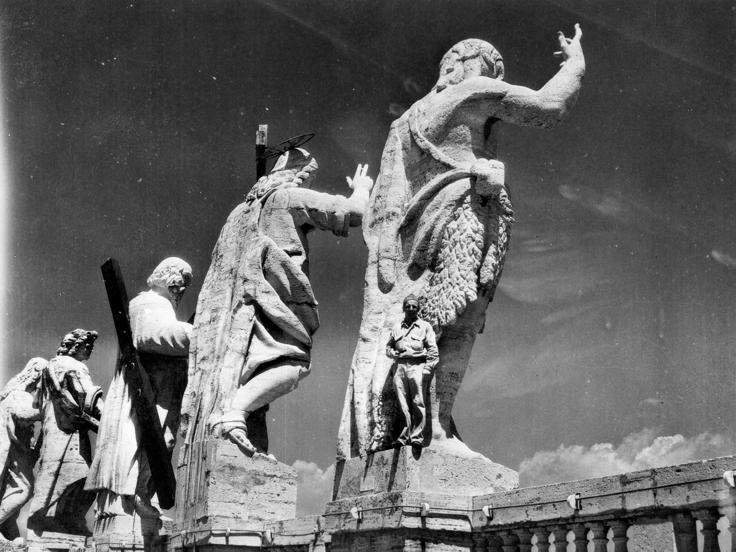 A historic black and white photo of 4 enormous sculptures seen from behind. On the plinth of the nearest sculpture stands Donald Beer, dwarfed by the scale of the figure above him.