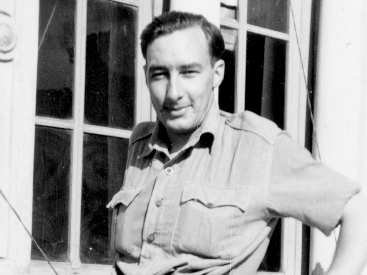 A historic black and white photo of a man in a uniform shirt. He is standing in front of a window and squinting into the sun.